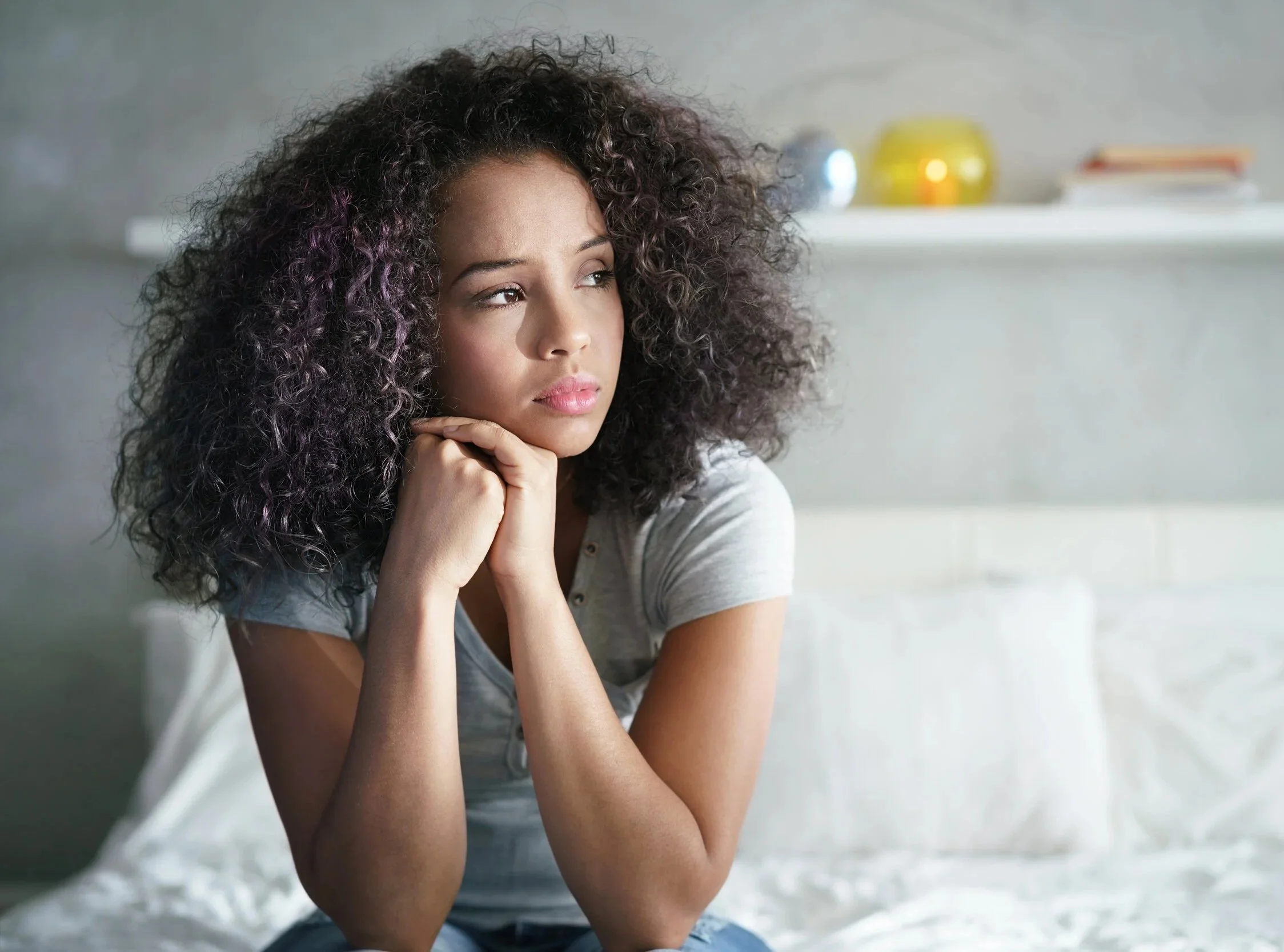 A young woman with curly hair sitting on a bed, looking thoughtful and pensive.