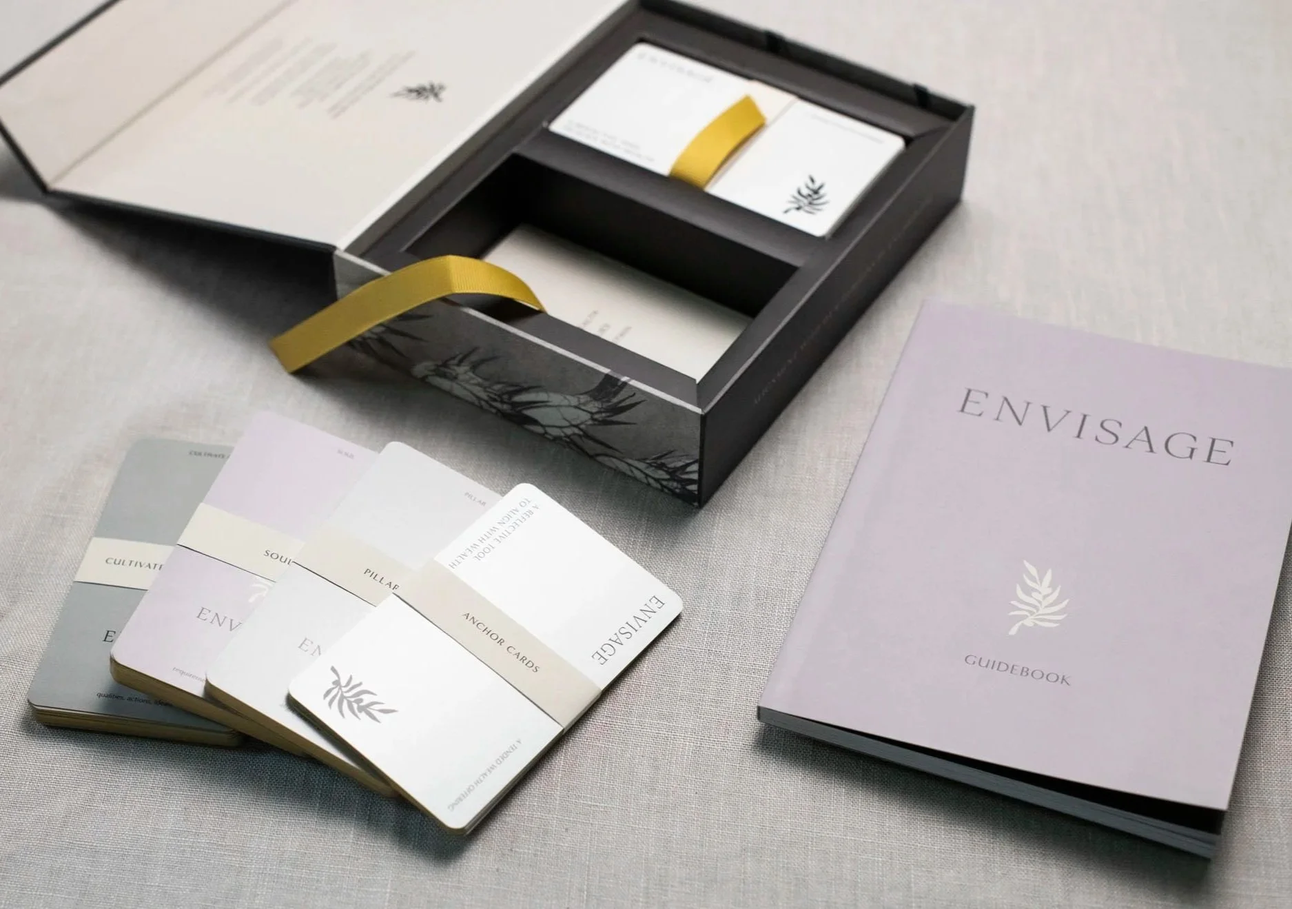ENVISAGE box open with 4 stacks of cards and guidebook resting on linen