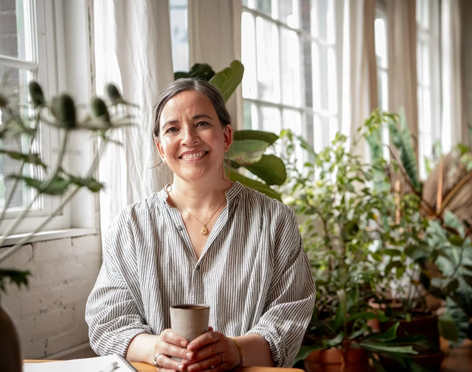 Megan deBoer smiling at camera holding a mug next to a light filled window with plants in the background