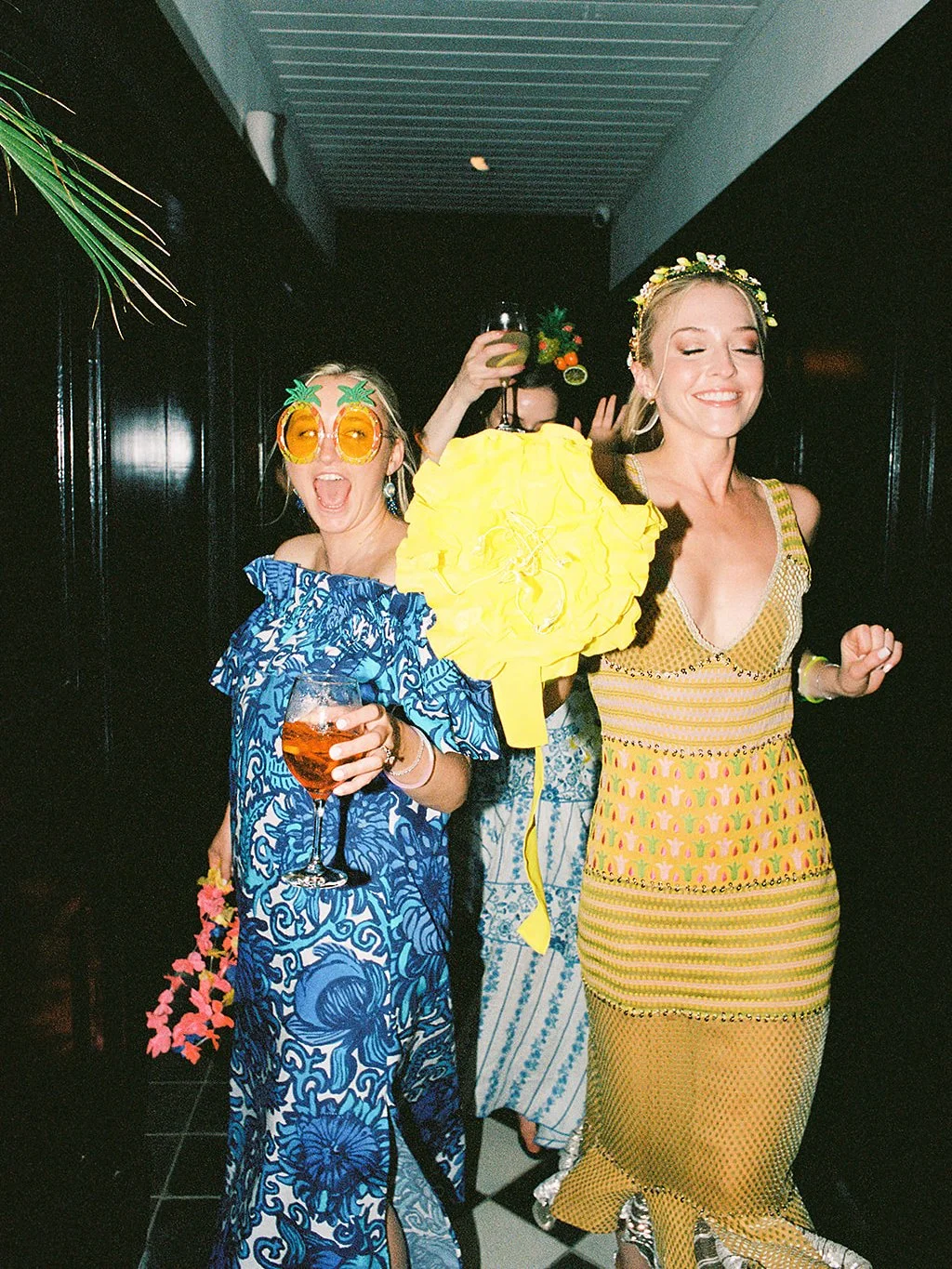 Guests in colorful dresses and pineapple sunglasses dance down a hallway with drinks, film photo