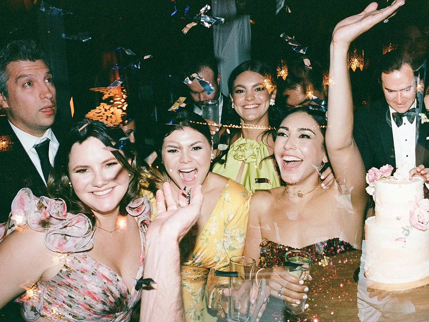Laughing bridesmaids and guests celebrate under falling confetti beside the wedding cake