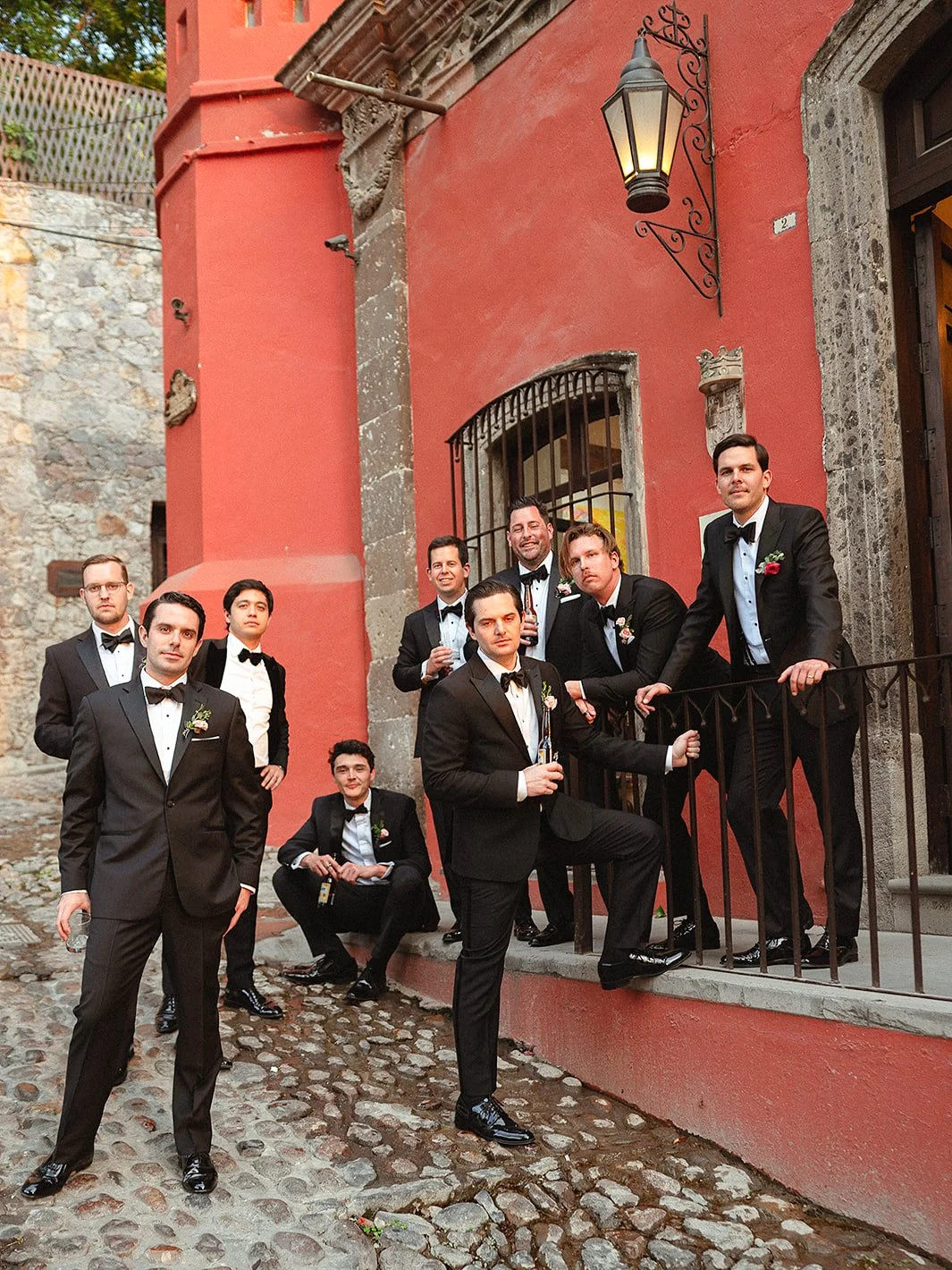 Groomsmen in tuxedos lean on a red colonial building on a cobblestone street in San Miguel de Allende, Mexico