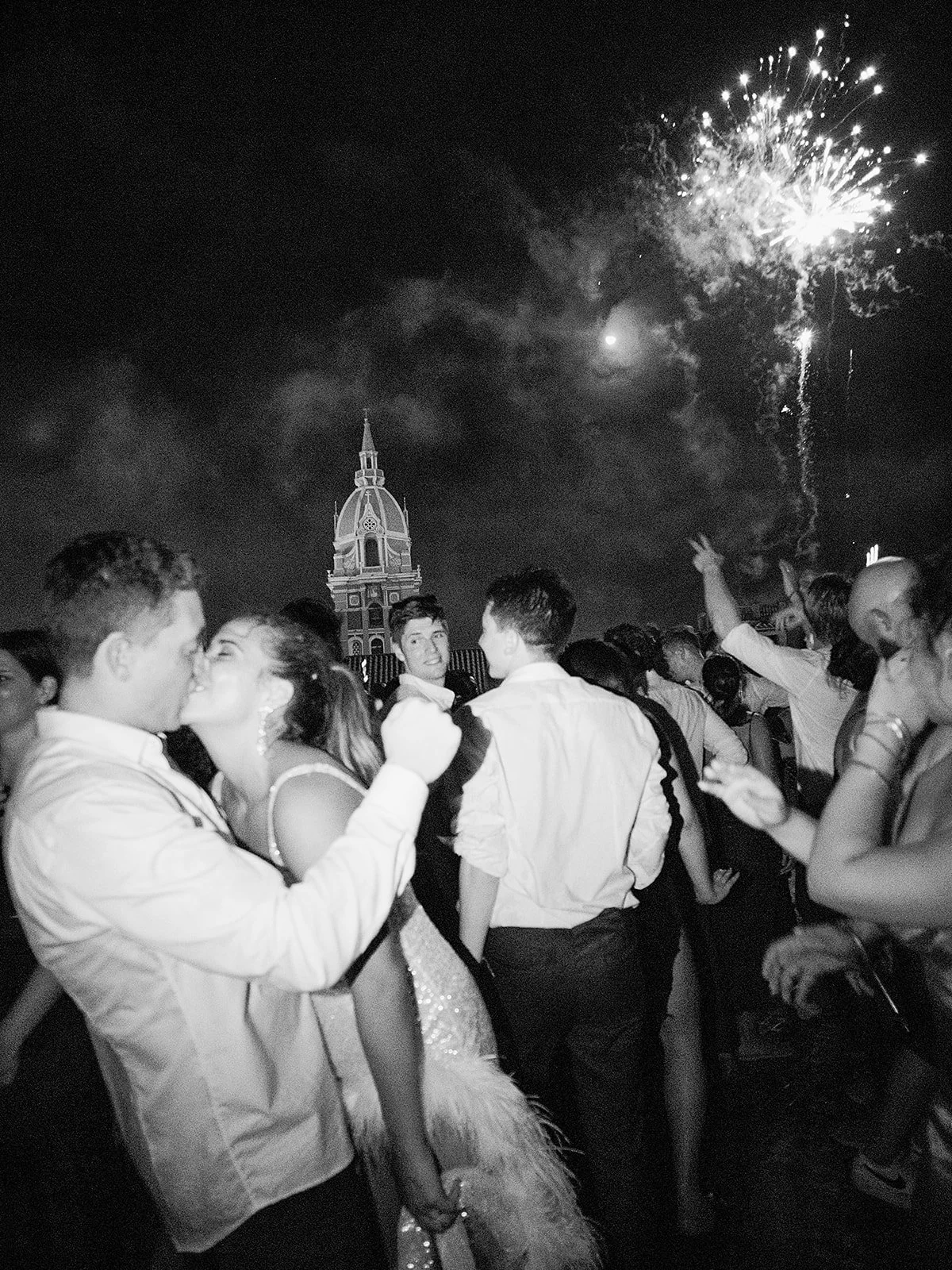 Black-and-white kiss on a rooftop dance floor as fireworks burst above a church dome