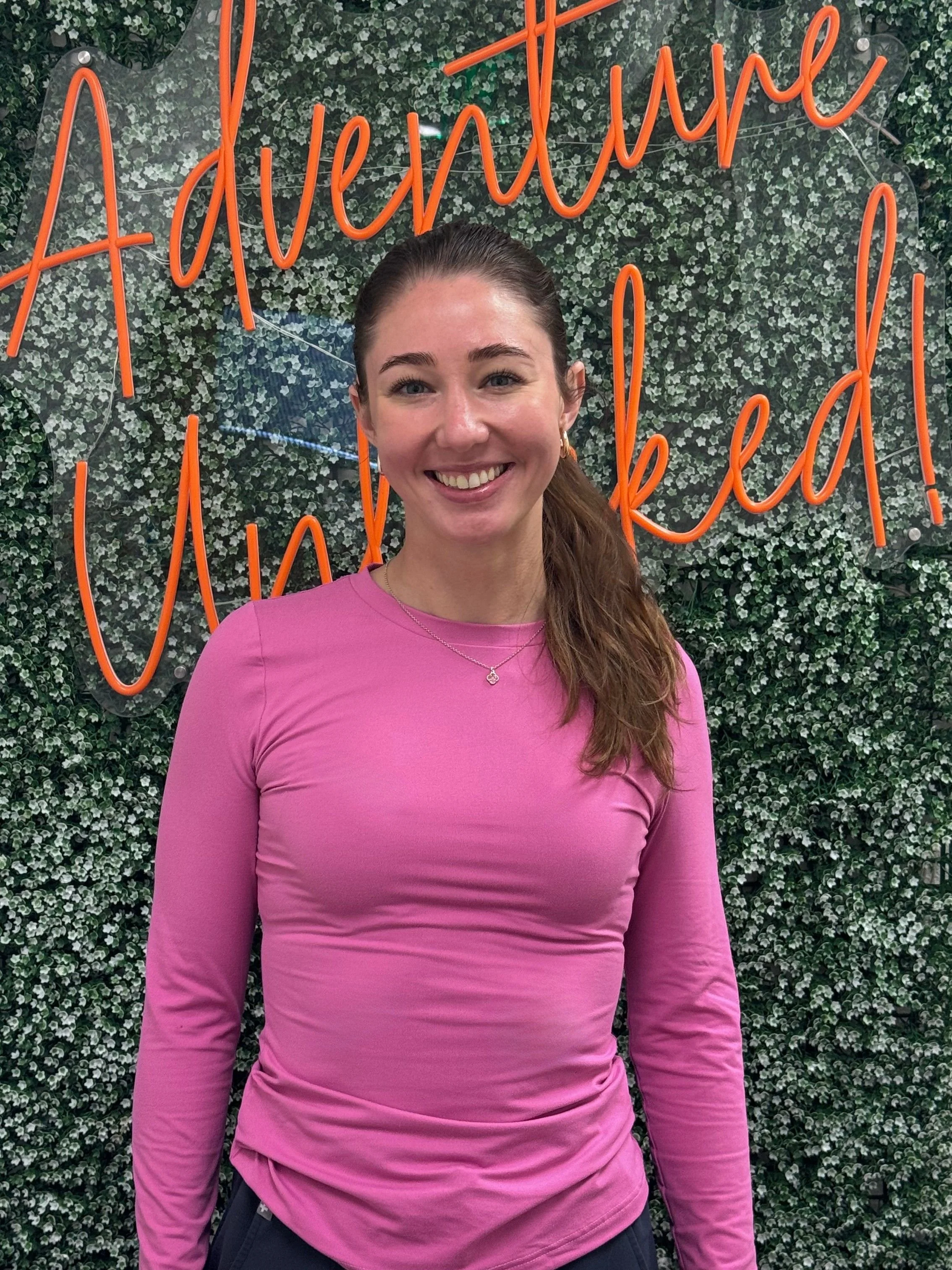 A young woman with long brown hair tied to the side, wearing a pink long-sleeve shirt and small earrings, standing in front of a wall with a neon sign that says 'Adventure Awaits' in orange cursive letters. She is smiling and has a necklace with a small pendant.