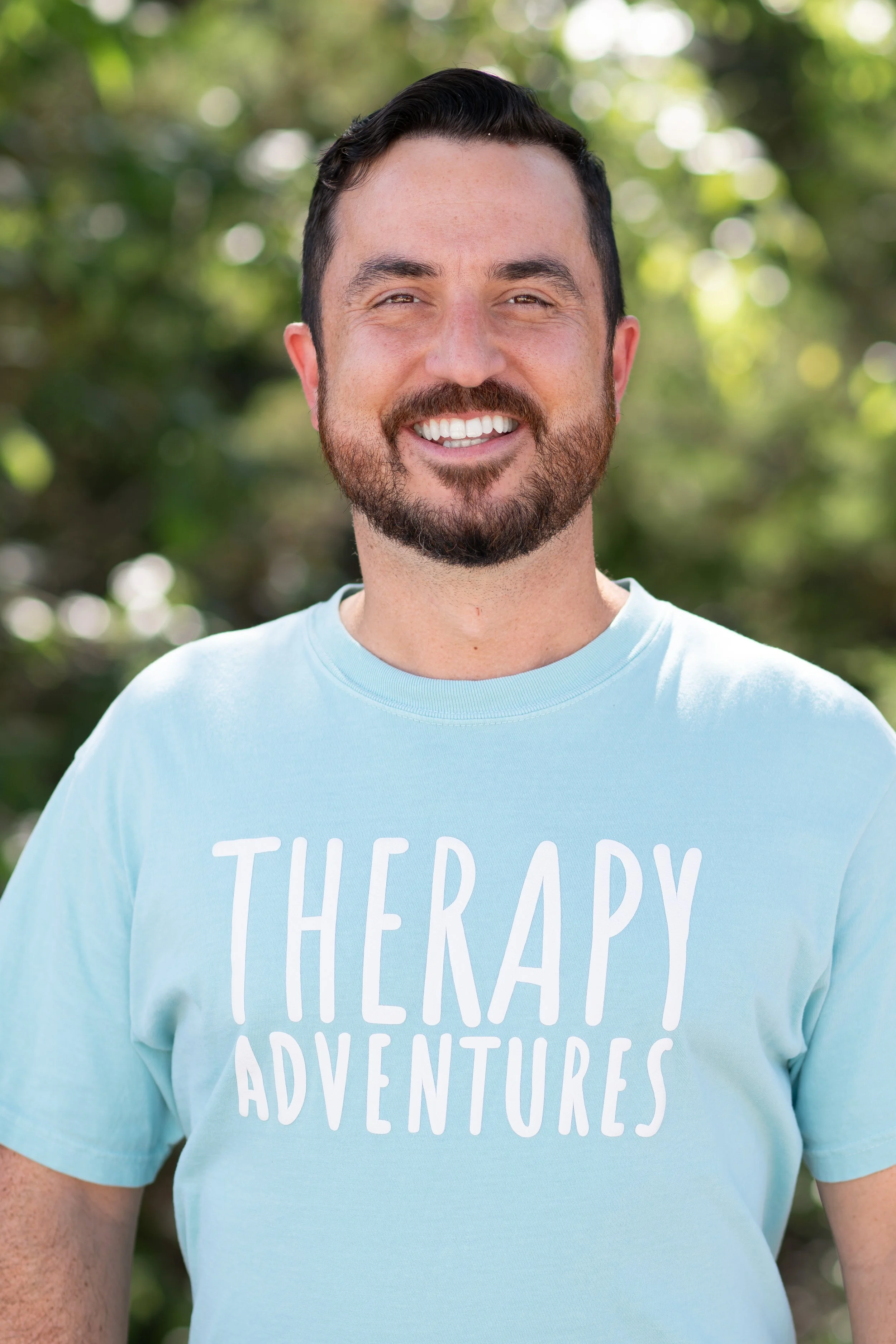 Man with dark hair, beard and mustache smiling against a background of trees