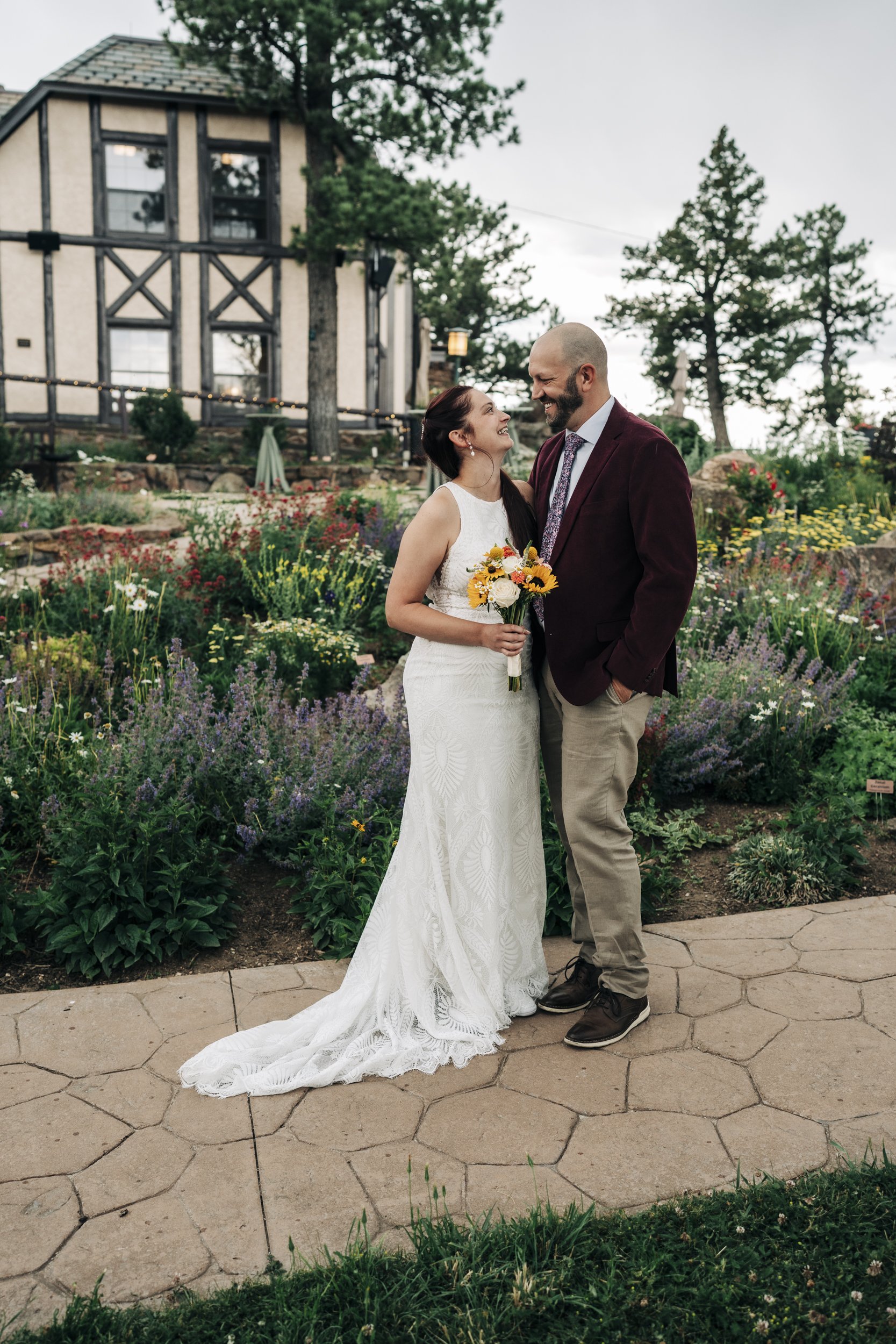 A bride and groom smiling and looking at each other outdoors in a garden filled with colorful flowers during a wedding. The bride is wearing a white lace wedding dress and holding a bouquet of yellow, orange, and white flowers. The groom is dressed i