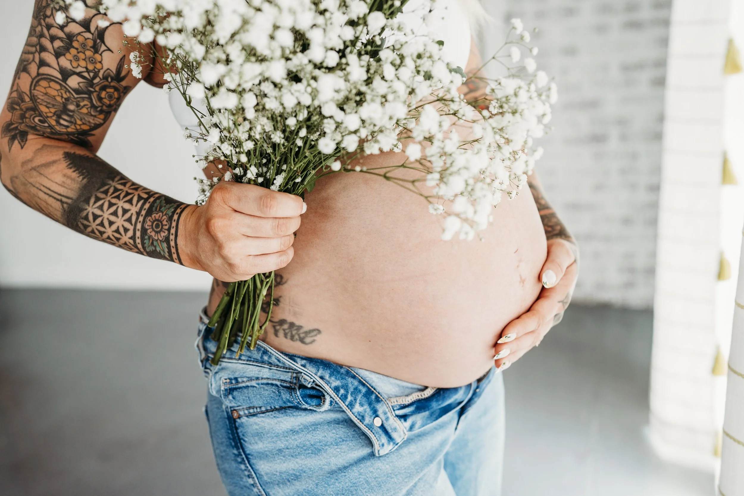 Pregnant woman holding a bouquet of white baby's breath flowers, with visible tattoos on her arm and side, wearing blue jeans, standing indoors near a window.