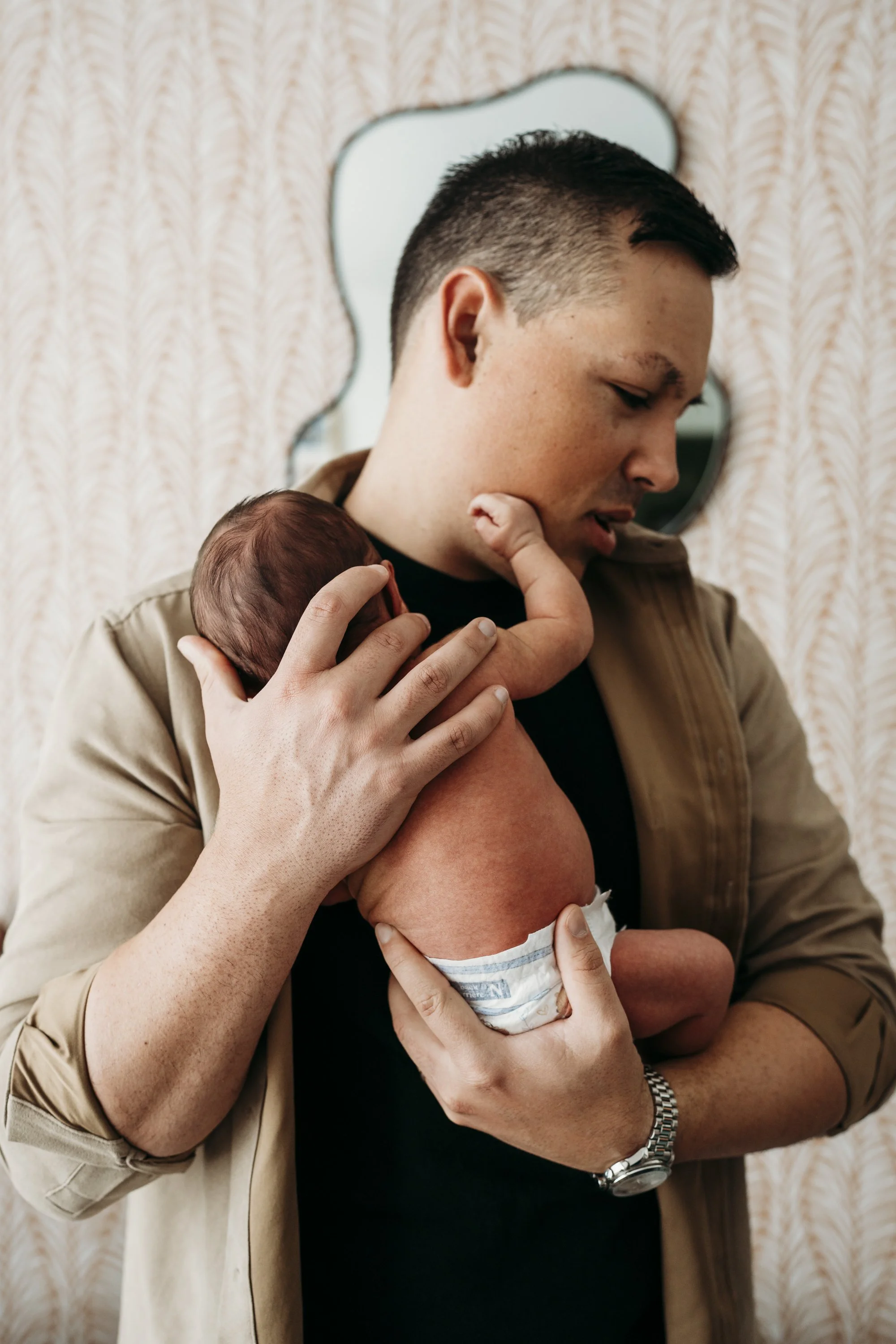 A man holding a newborn baby close to his chest, with the baby's face resting on his shoulder, indoors with patterned wallpaper and a mirror in the background.