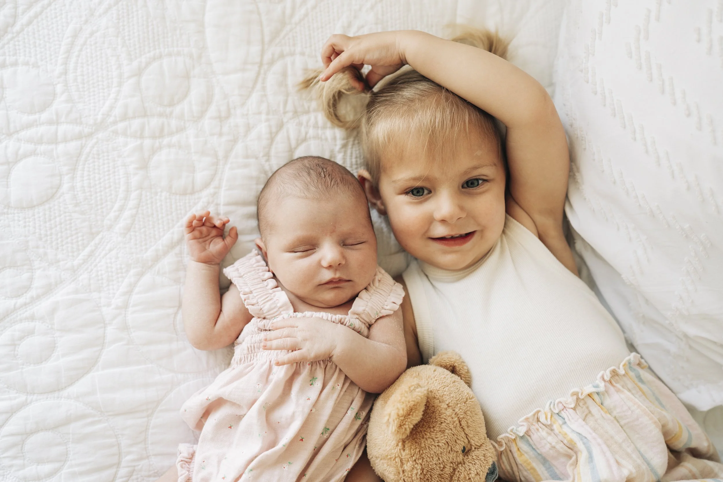 Two young children, one asleep and a toddler awake, lying on a white quilted bed. The toddler has blonde hair, blue eyes, and is holding a plush teddy bear while smiling. The baby has a little hair, closed eyes, and is dressed in a light pink dress.