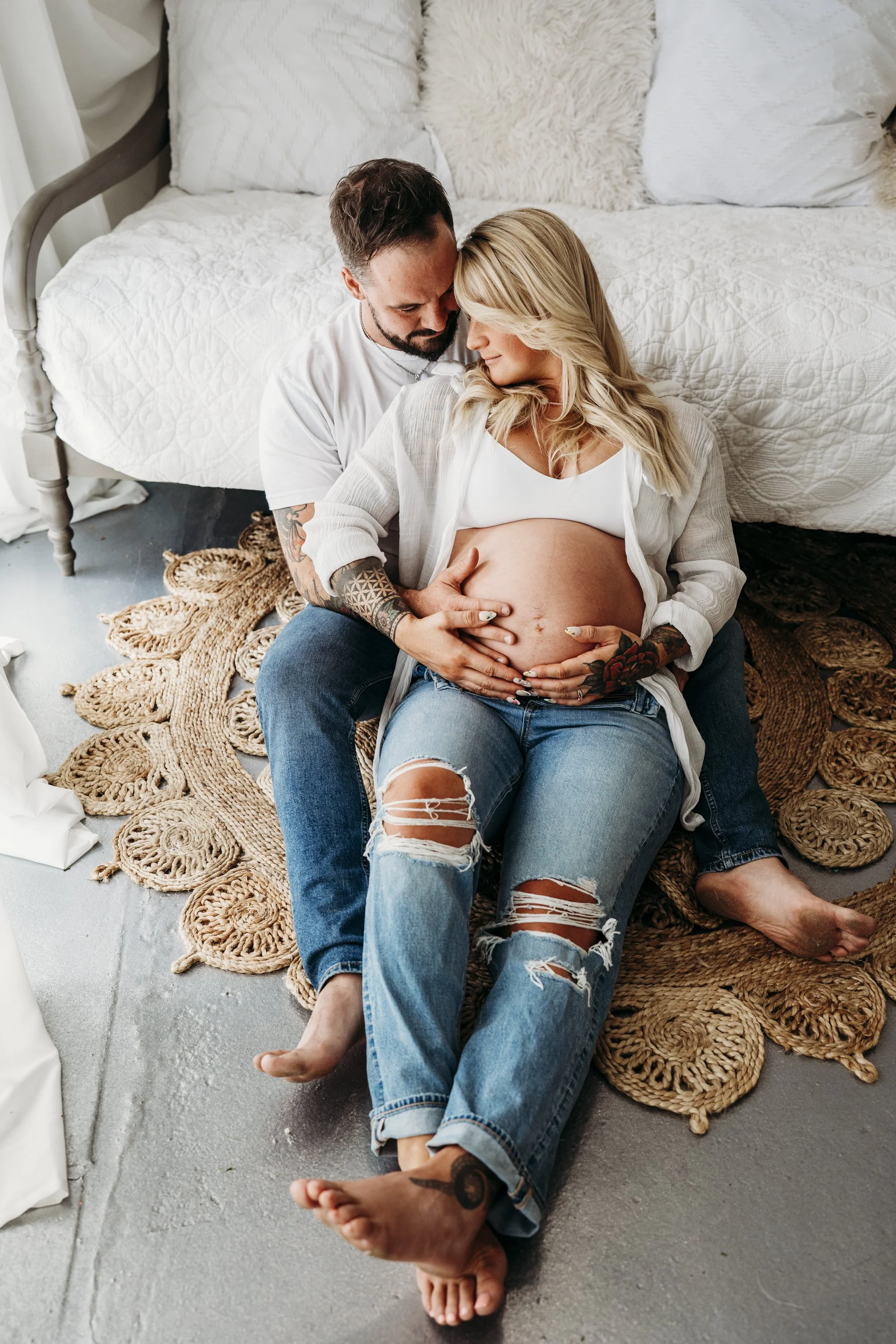 A couple sitting on the floor, embracing, with the woman's pregnant belly visible, in a cozy indoor setting.