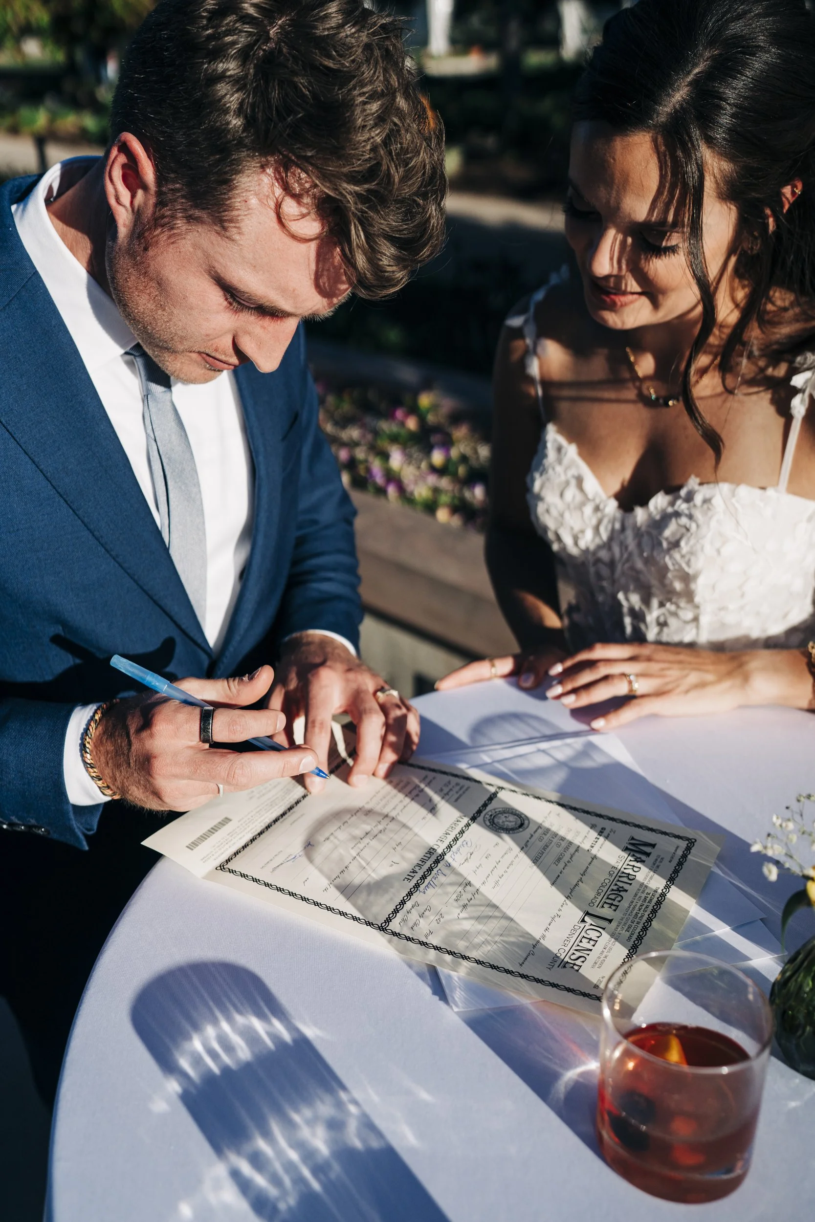 A couple signing a marriage license outdoors, with a glass of red wine on the table and flowers in the background.