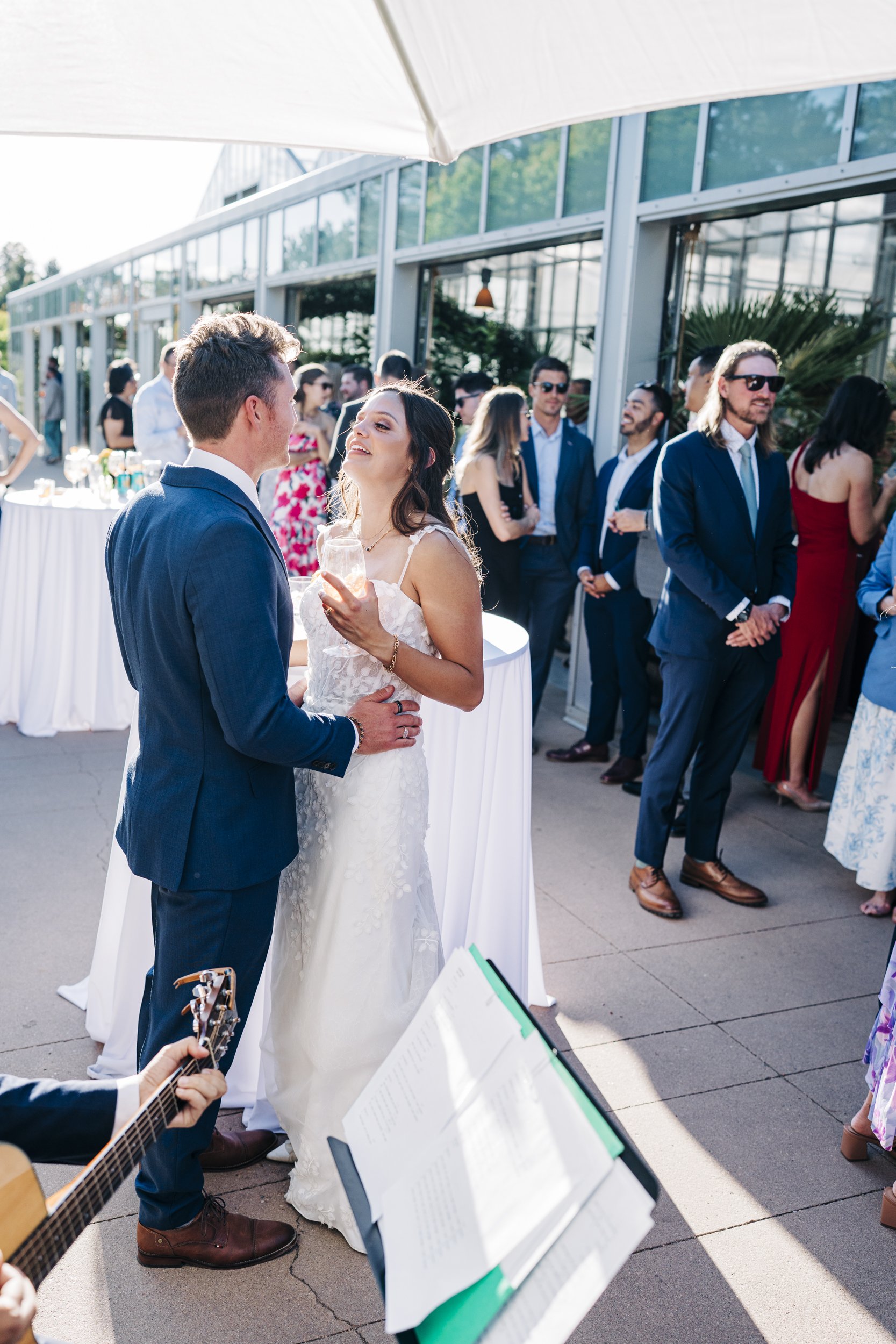 A bride and groom celebrating their wedding outdoors, surrounded by guests in formal attire, with a musician playing guitar in the foreground.