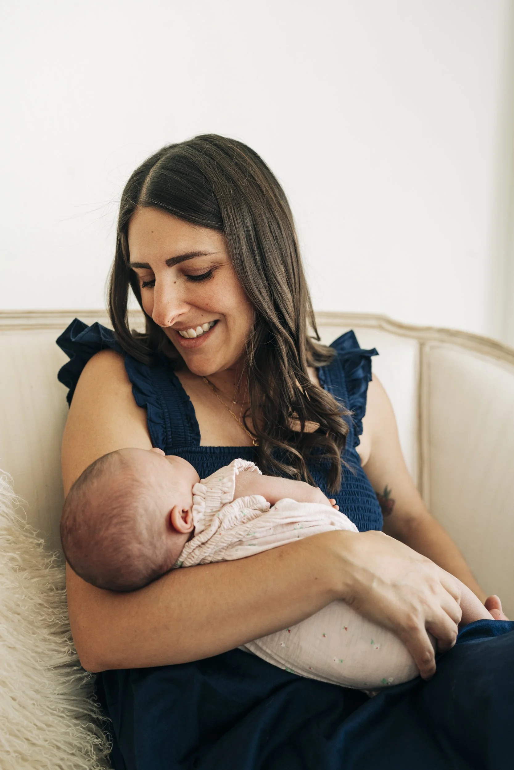 A woman with dark hair wearing a blue top sitting on a cream-colored couch holding a baby, looking down and smiling.