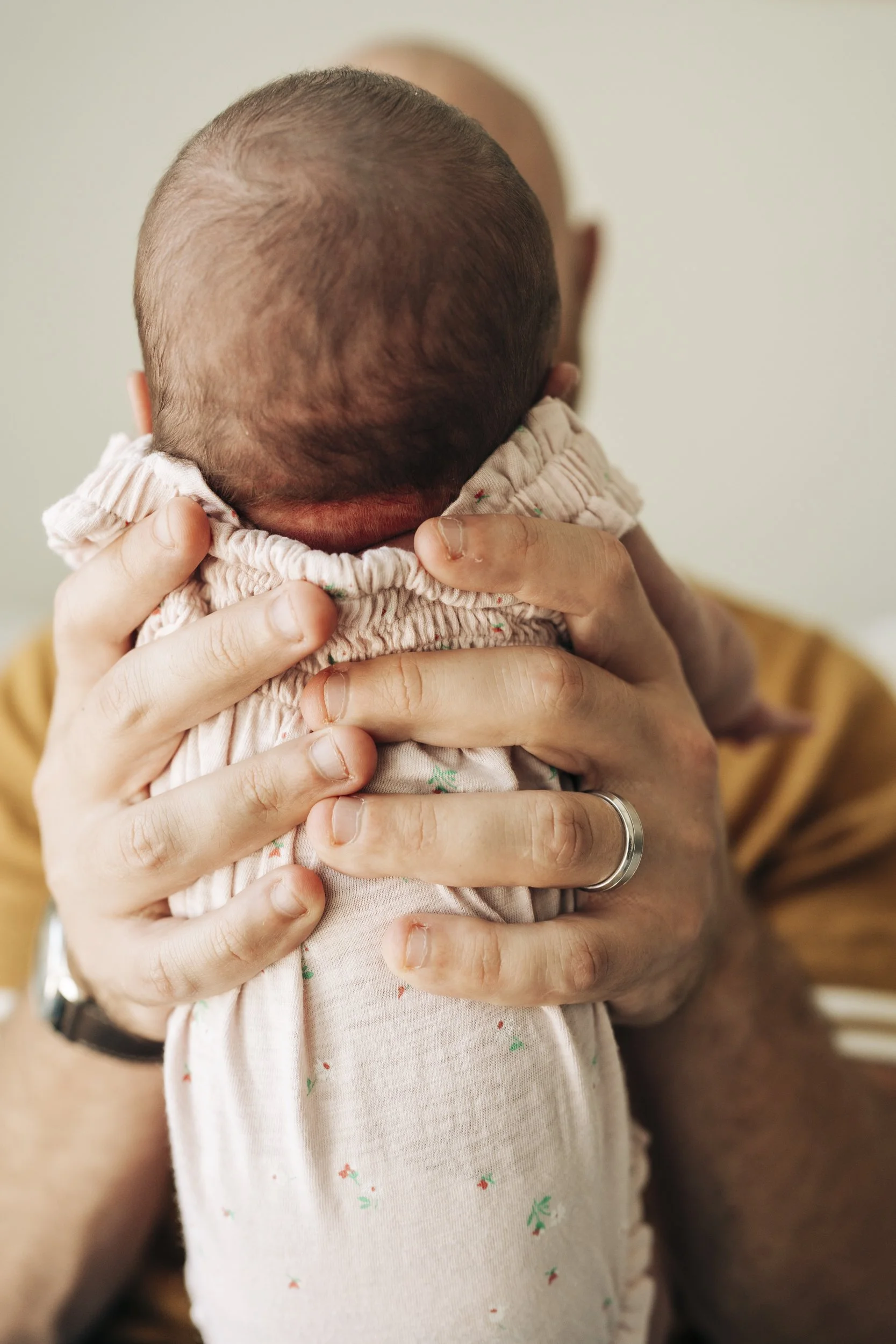 A person holding a baby close to their face, viewed from behind, with focus on the back of the baby's head and the person's hands. The baby is wearing a light-colored, patterned outfit and has a bald or very short hair. The person holding the baby is