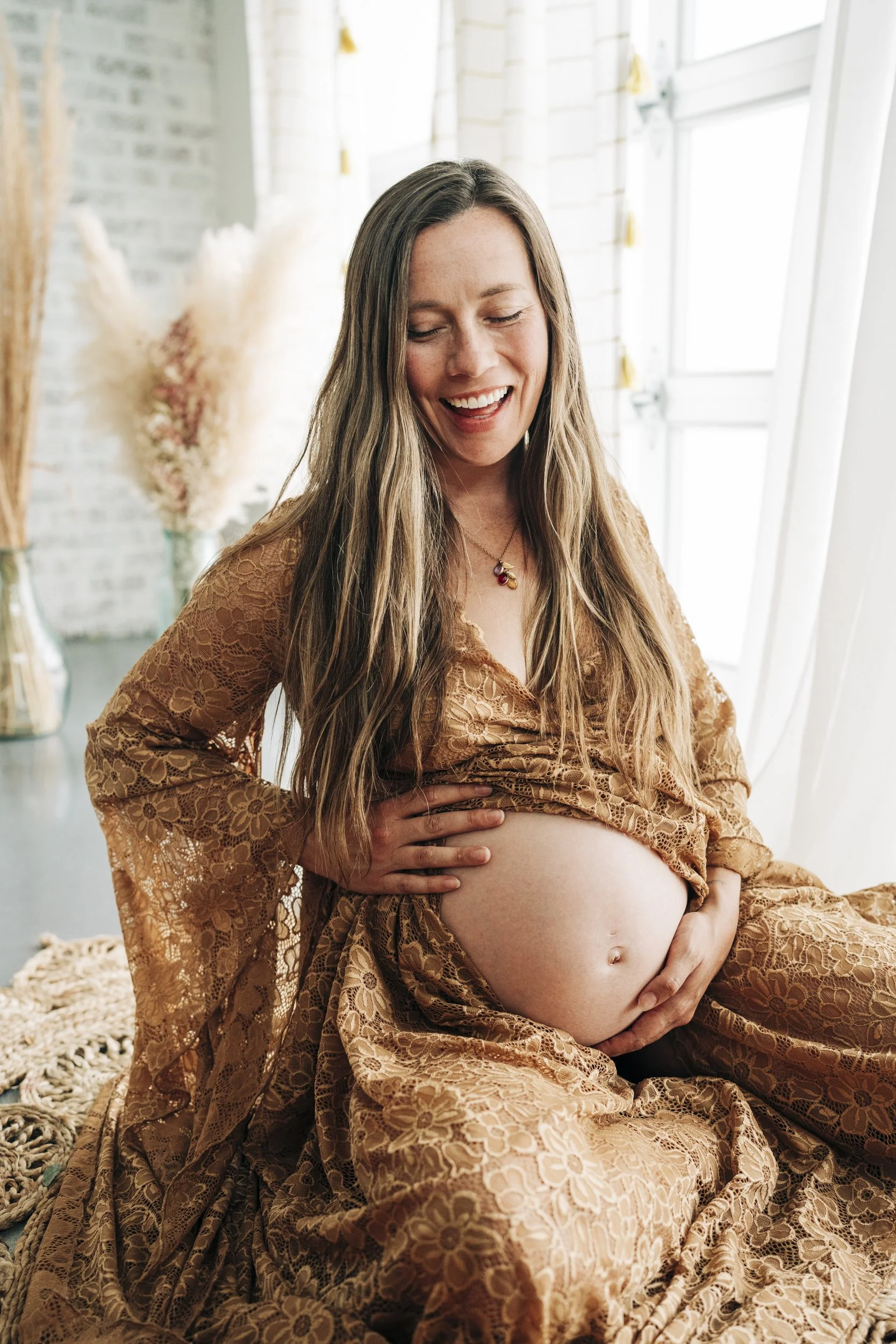 Pregnant woman in a brown lace dress sitting on a woven rug in a bright room, smiling and holding her belly.