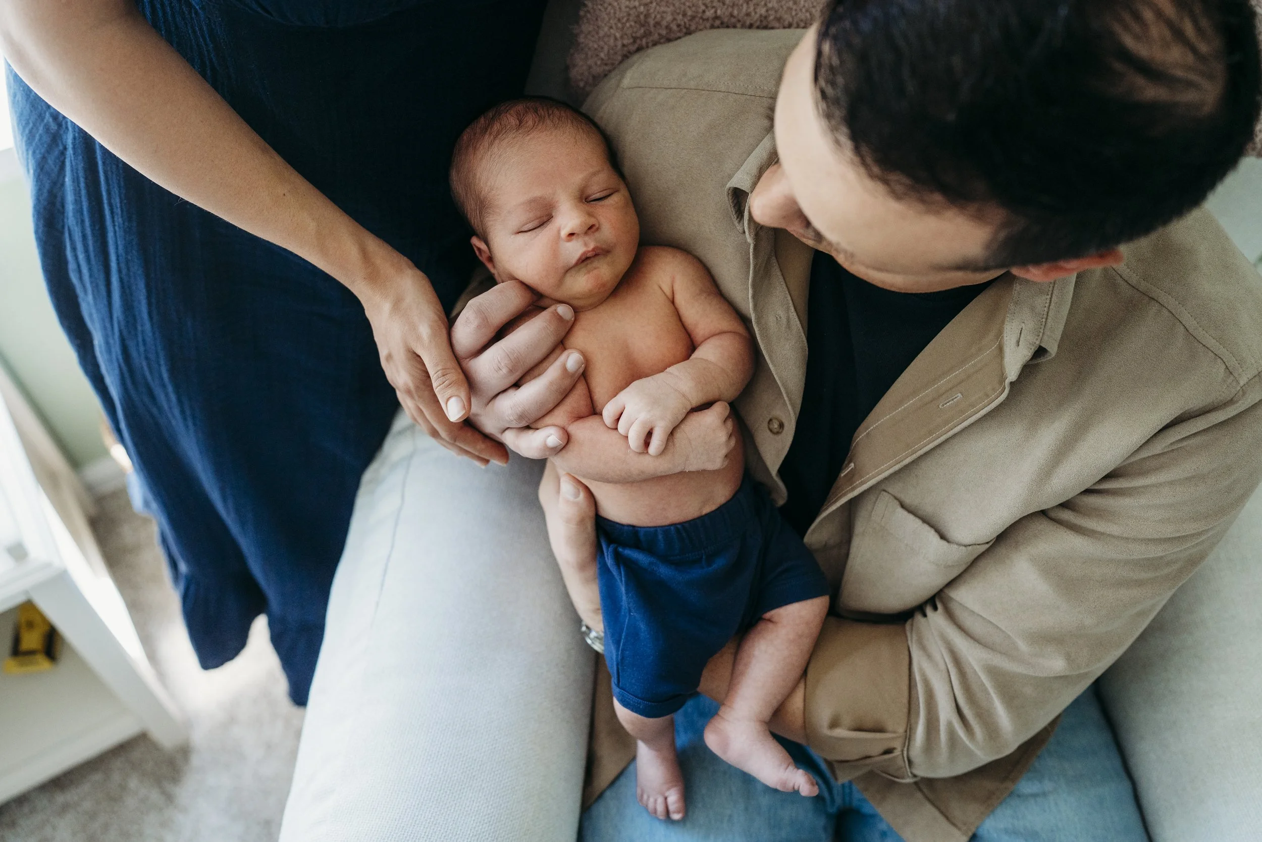 A man holding a sleeping baby with a woman's hand gently touching the baby's head, sitting on a beige couch in a cozy room with a window in the background.
