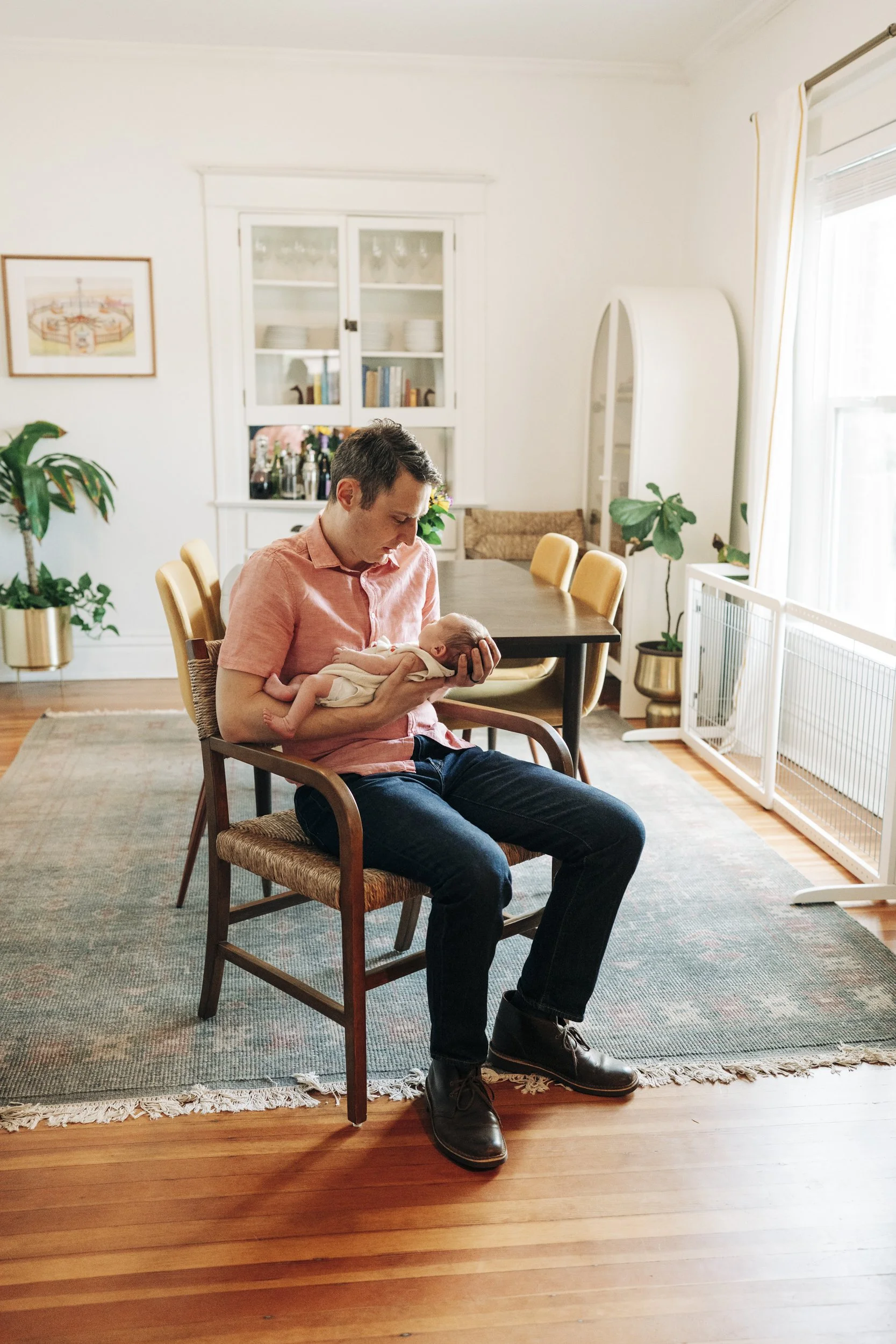 A man in a pink shirt and dark jeans holding a newborn baby in a white outfit inside a bright, cozy living room with wooden floors, green plants, a dining table, and large windows.