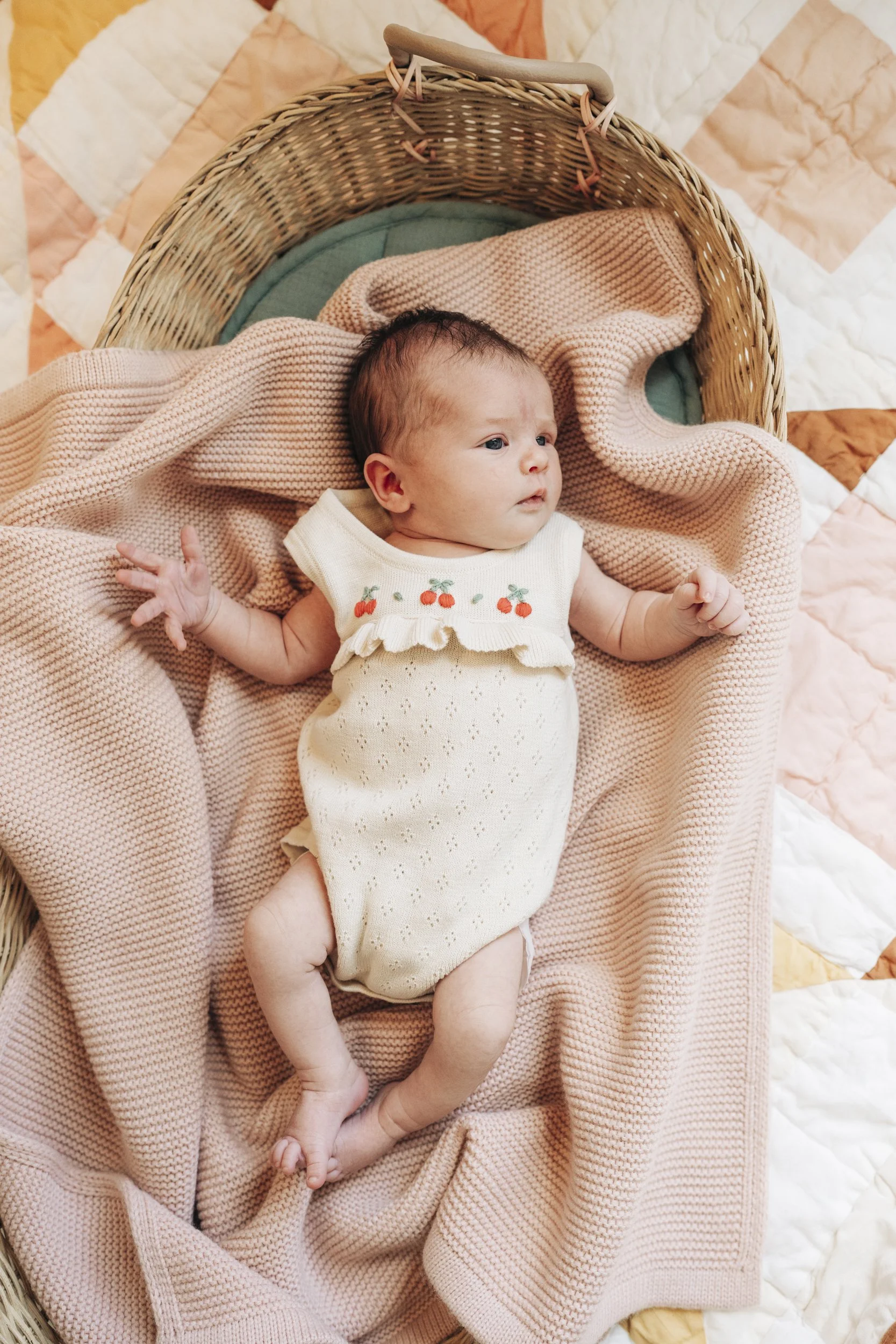 A baby lying in a woven bassinet with peach, green, and white bedding. The baby is wearing a cream-colored outfit with embroidered cherries and a ruffled collar, surrounded by soft blankets.