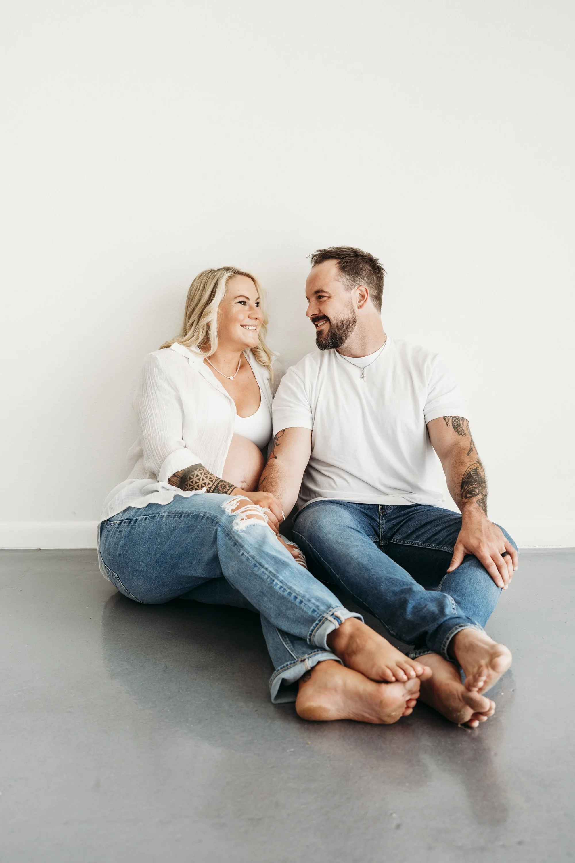 A pregnant woman and a man sitting on the floor, smiling and looking at each other. The woman is cradling her belly and they are both in casual white shirts and jeans, with a plain white wall in the background.