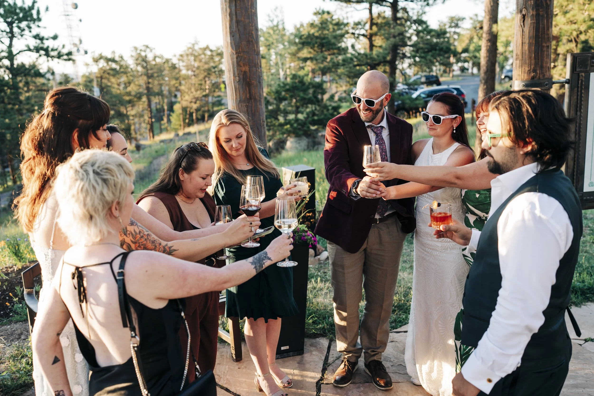 A group of people at an outdoor gathering, with some dressed in formal attire, raising glasses for a toast in a forested area during sunset.