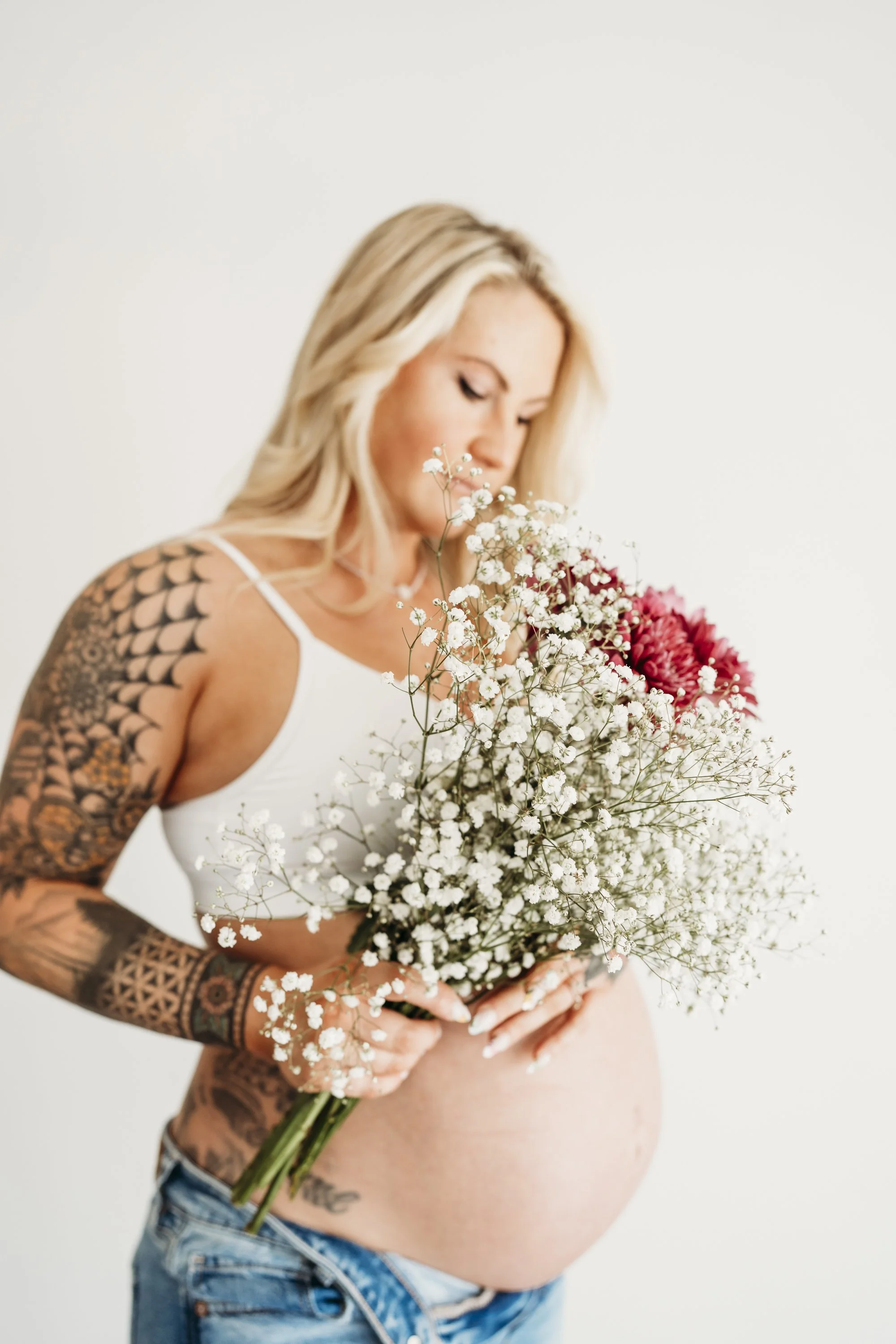 Pregnant woman with tattoos on arms holding a bouquet of white and pink flowers.