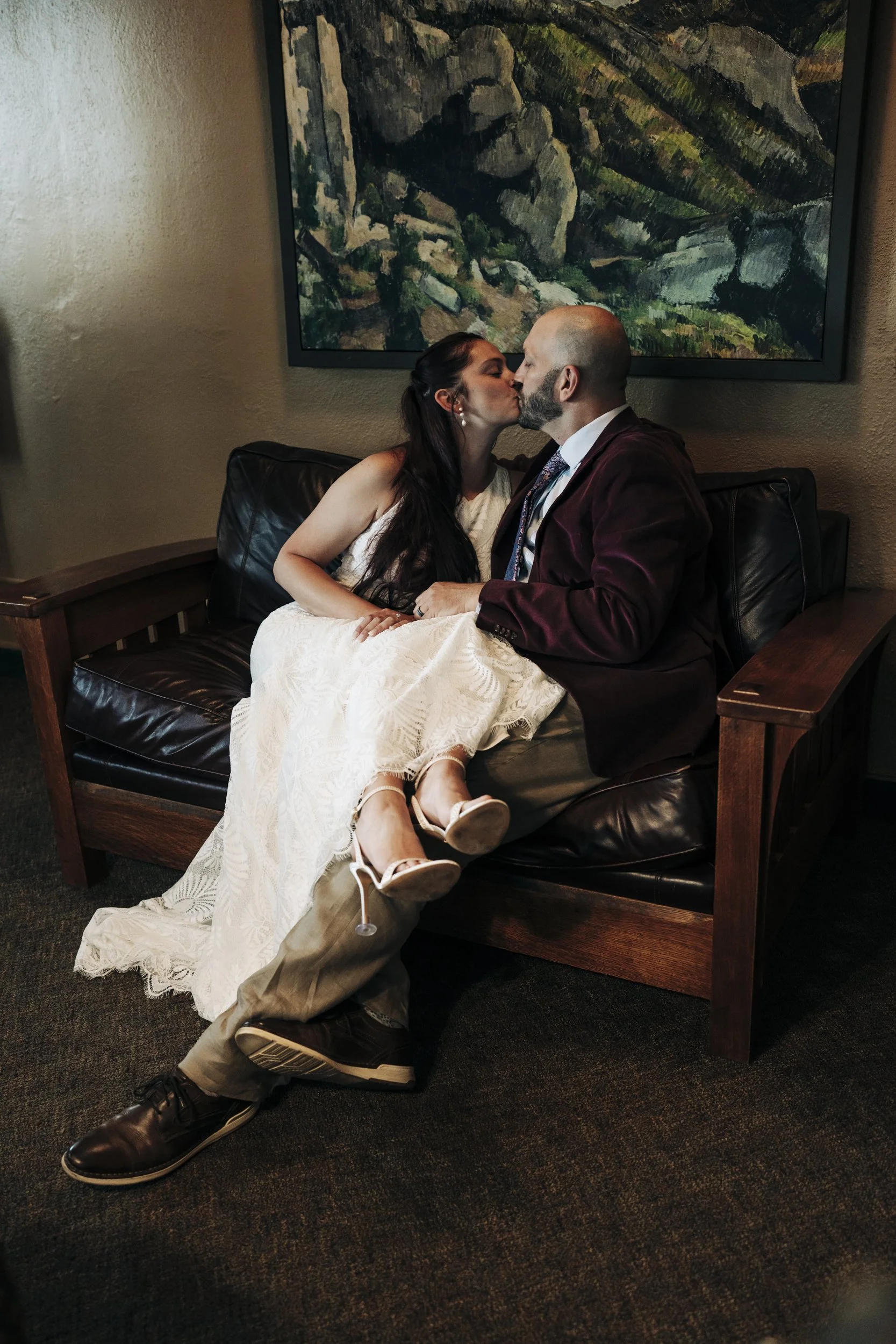 A man in a suit with a beard and a woman in a wedding dress are sitting on a black leather and wood bench, sharing a kiss in a room with a large landscape painting hanging on the wall behind them.