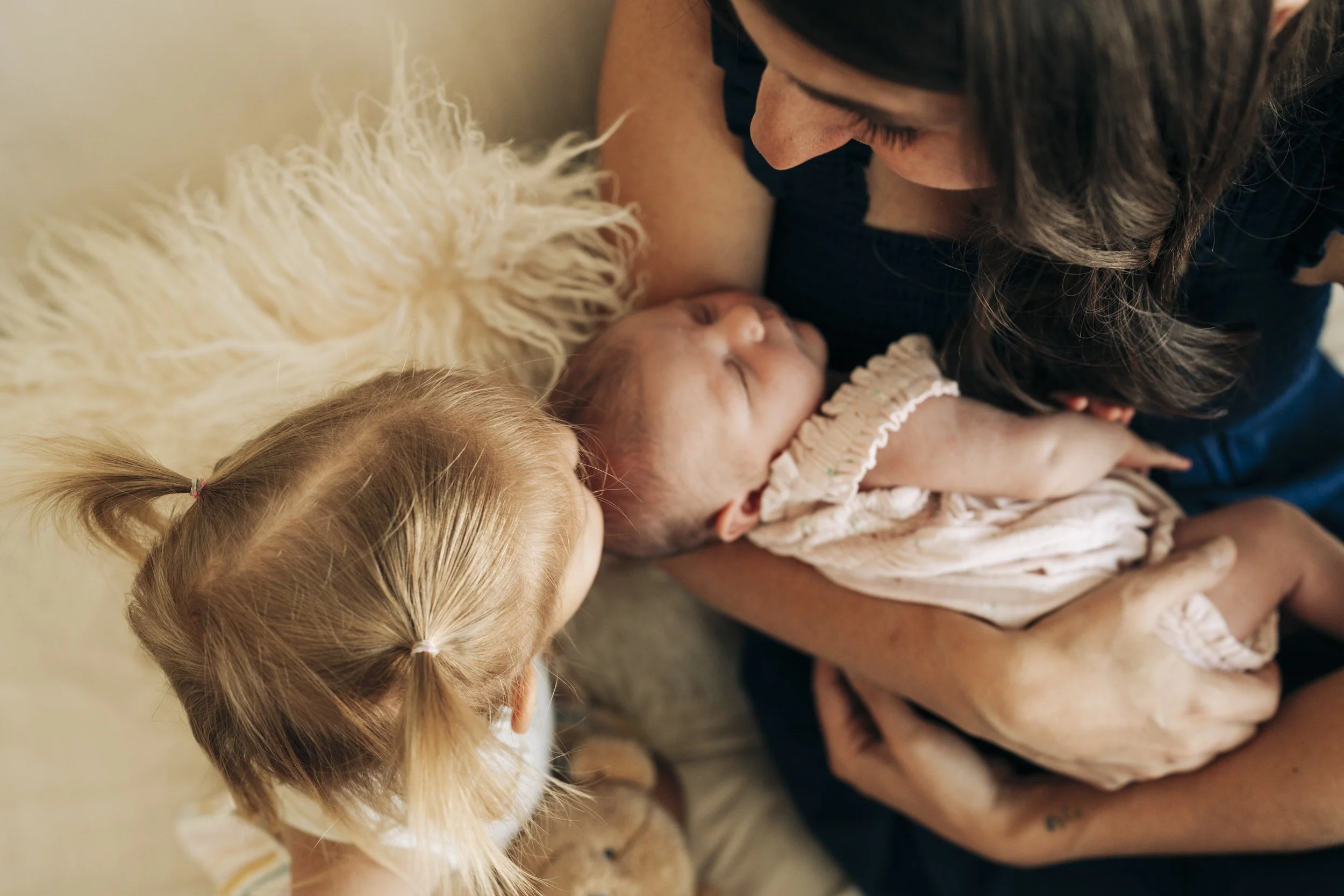 A woman holding a sleeping newborn baby in her arms, with a young girl with blonde hair and pigtails looking at the baby, lying on a soft blanket.