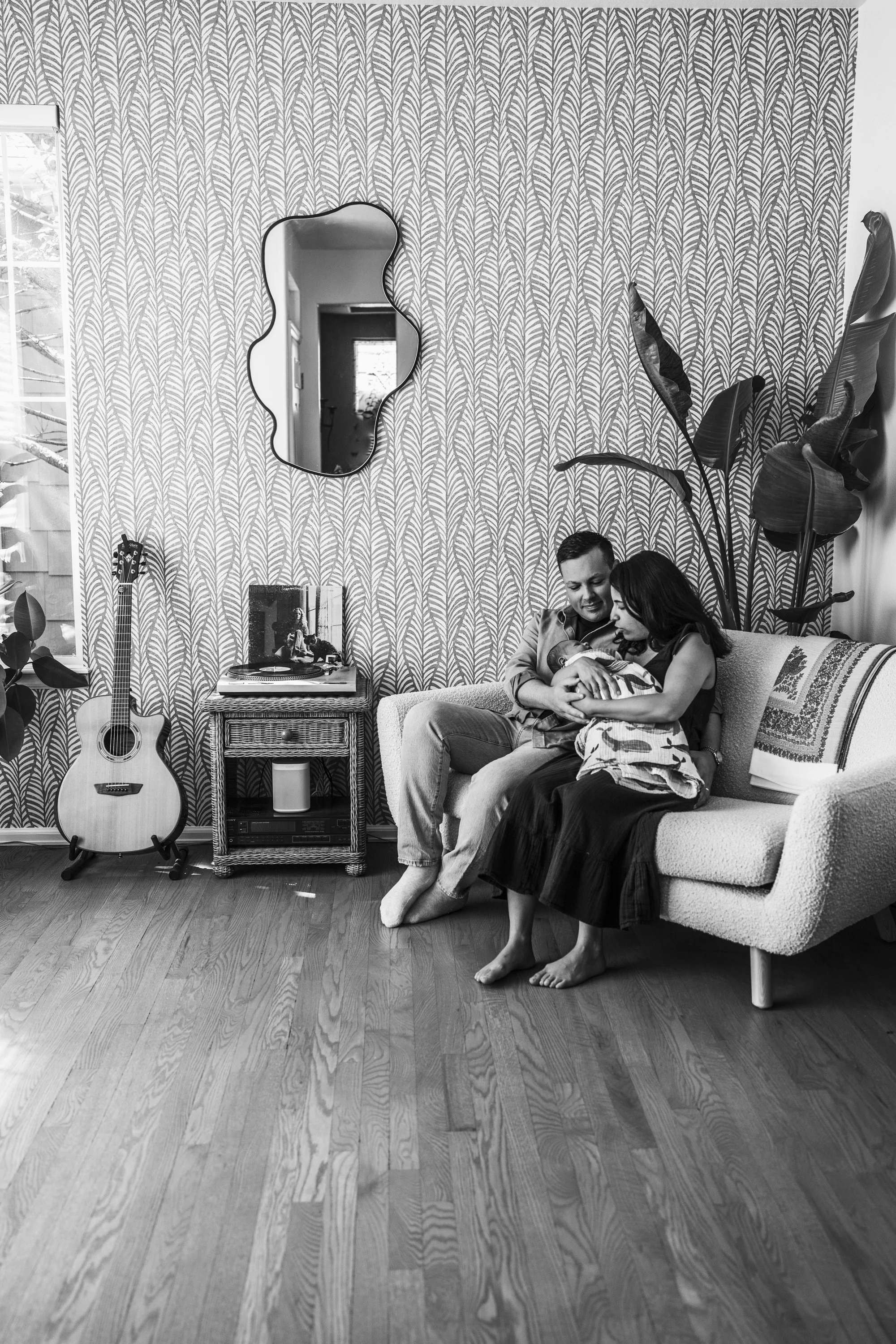 A black-and-white photo of a couple sitting on a couch, holding a newborn baby, in a cozy living room with patterned wallpaper, a guitar on the floor, a side table with records, and a large plant.