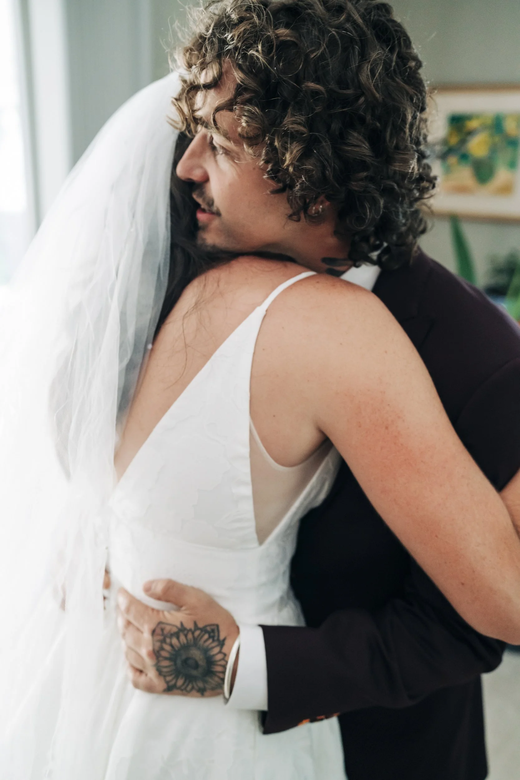 A close-up of a couple hugging, with the man wearing a dark suit and the woman in a white dress. The man has curly hair and a tattoo on his hand. The woman has dark hair and is wearing a veil, suggesting it may be a wedding.