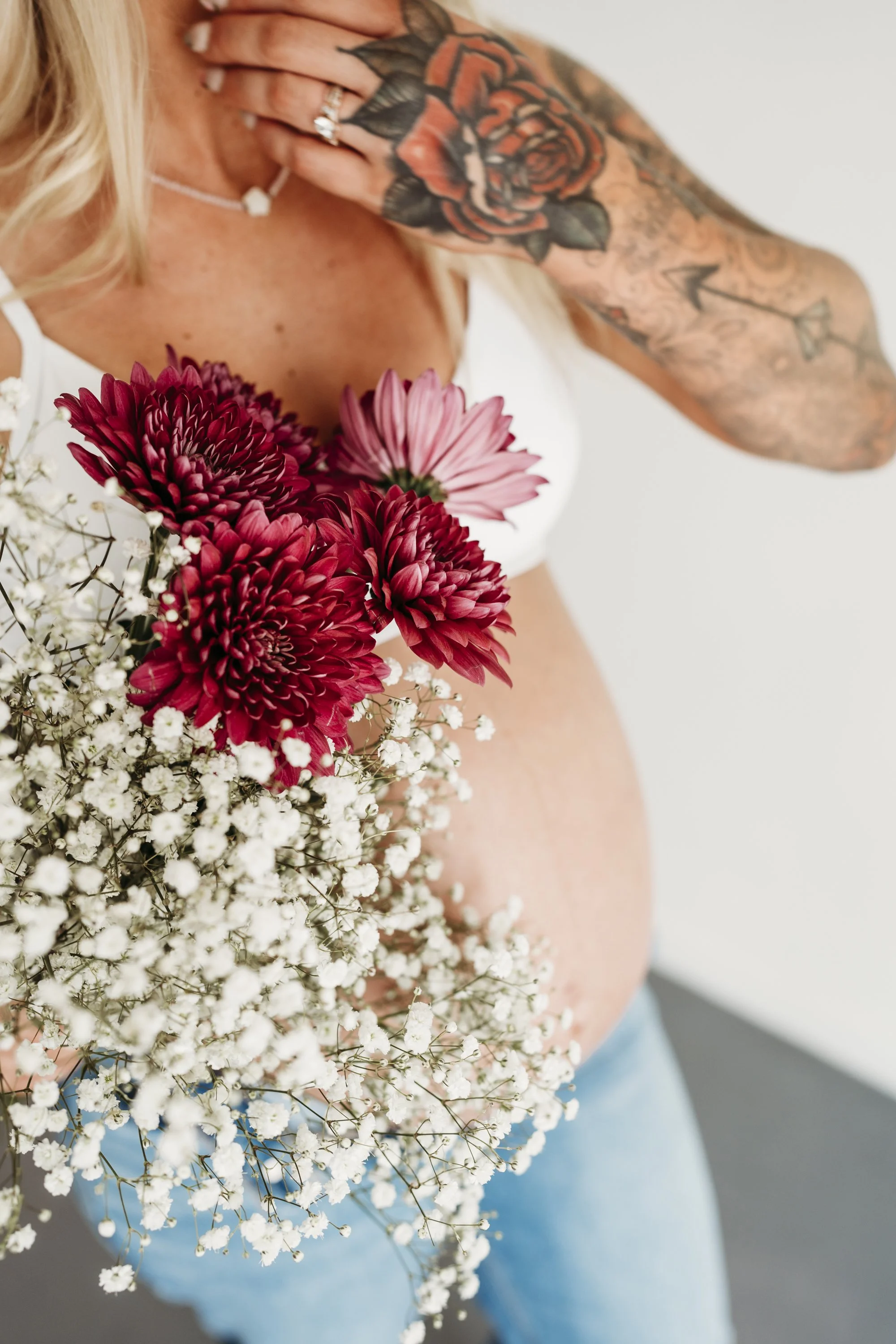 Woman holding a bouquet of pink and white flowers, with visible tattoos on her arm, wearing a white top and blue jeans.