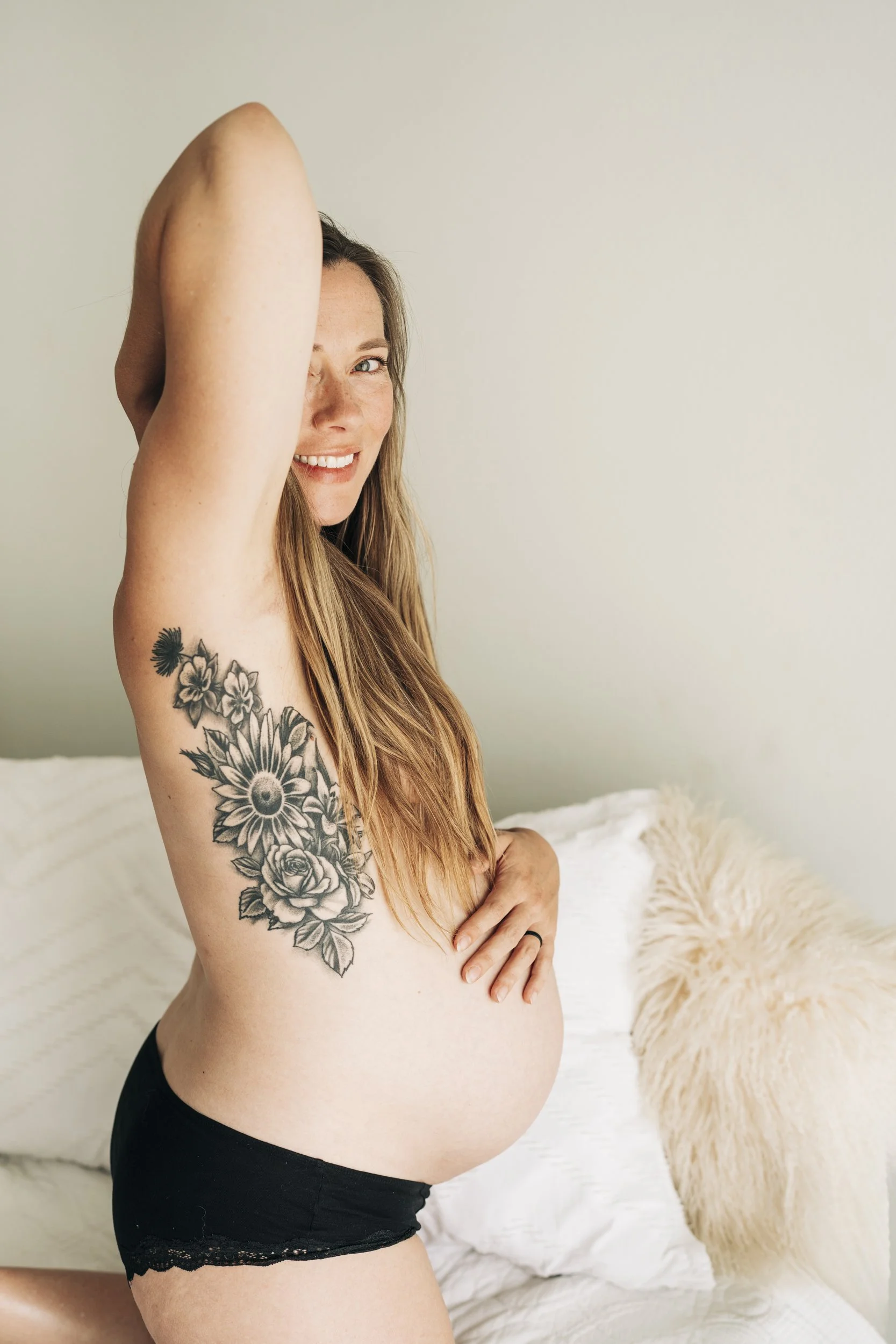 A pregnant woman with long hair, smiling, with a floral tattoo on her arm, wearing black shorts, sitting on a bed with white bedding and a fluffy pillow.