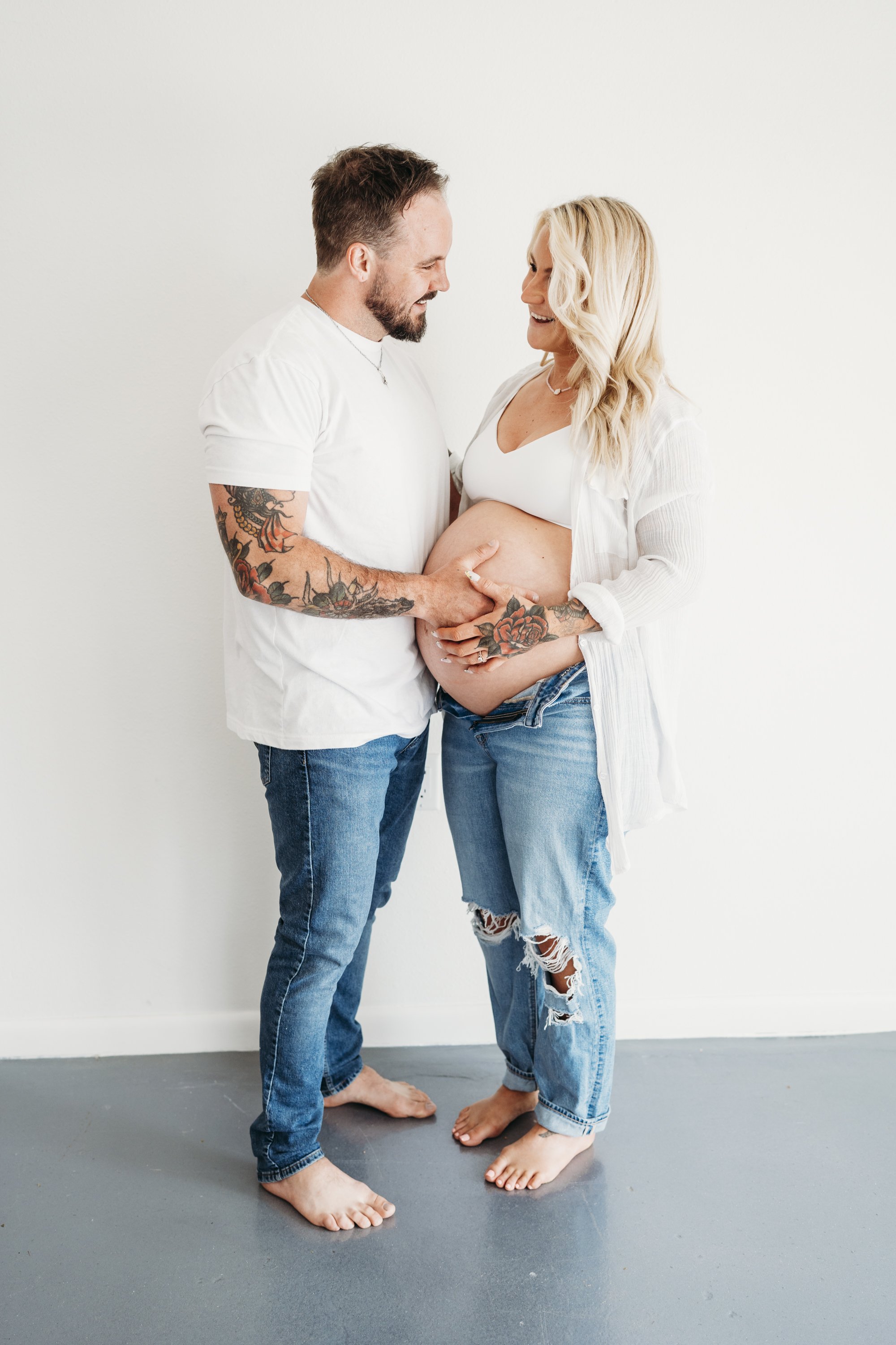 A pregnant woman and a man are standing together, touching her belly and smiling at each other in a bright, plain room.