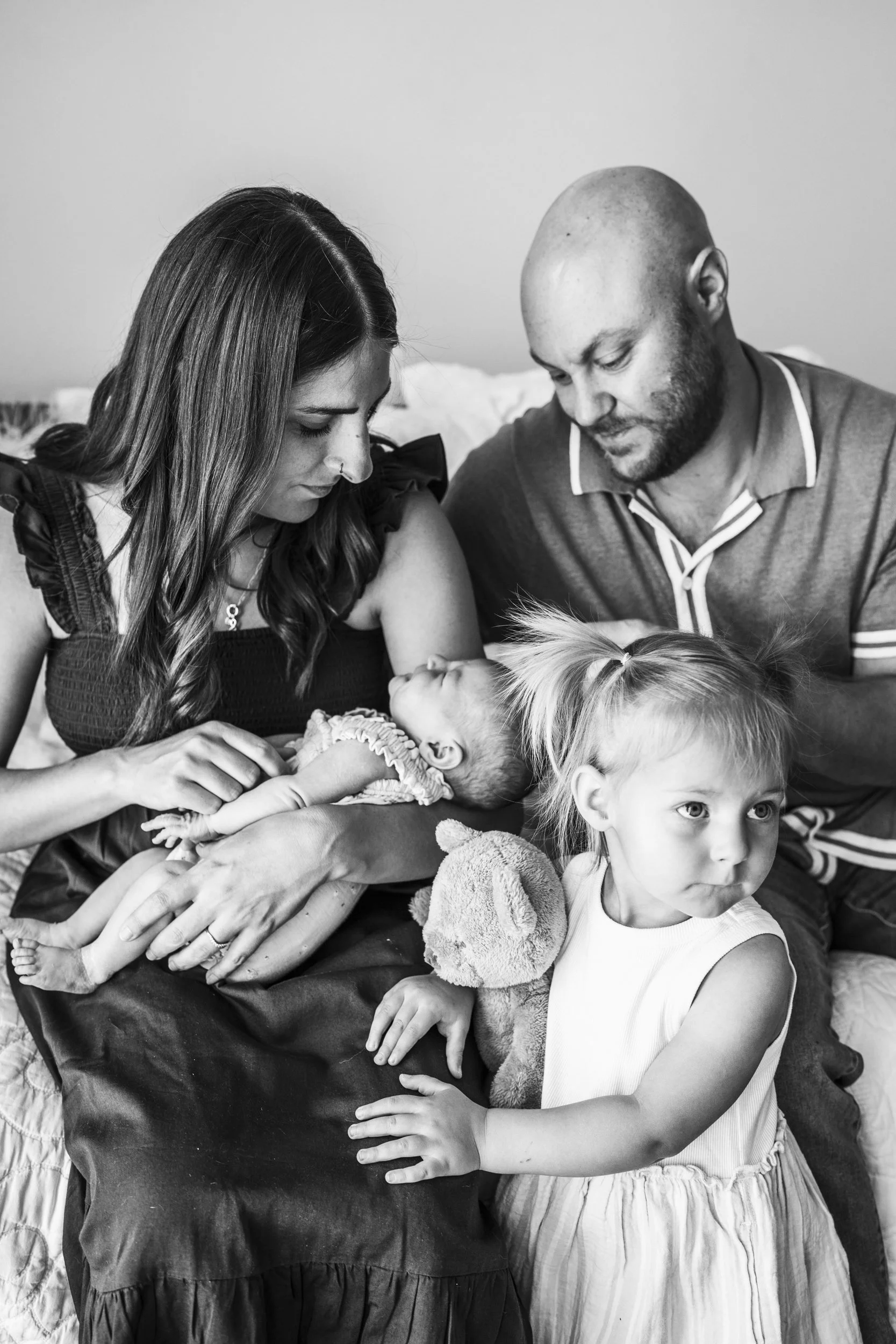 A black-and-white photo of a family with a newborn, a young girl, and two adults sitting together on a bed. The mother holds the baby, the father looks at the baby, and the young girl holds a stuffed animal.