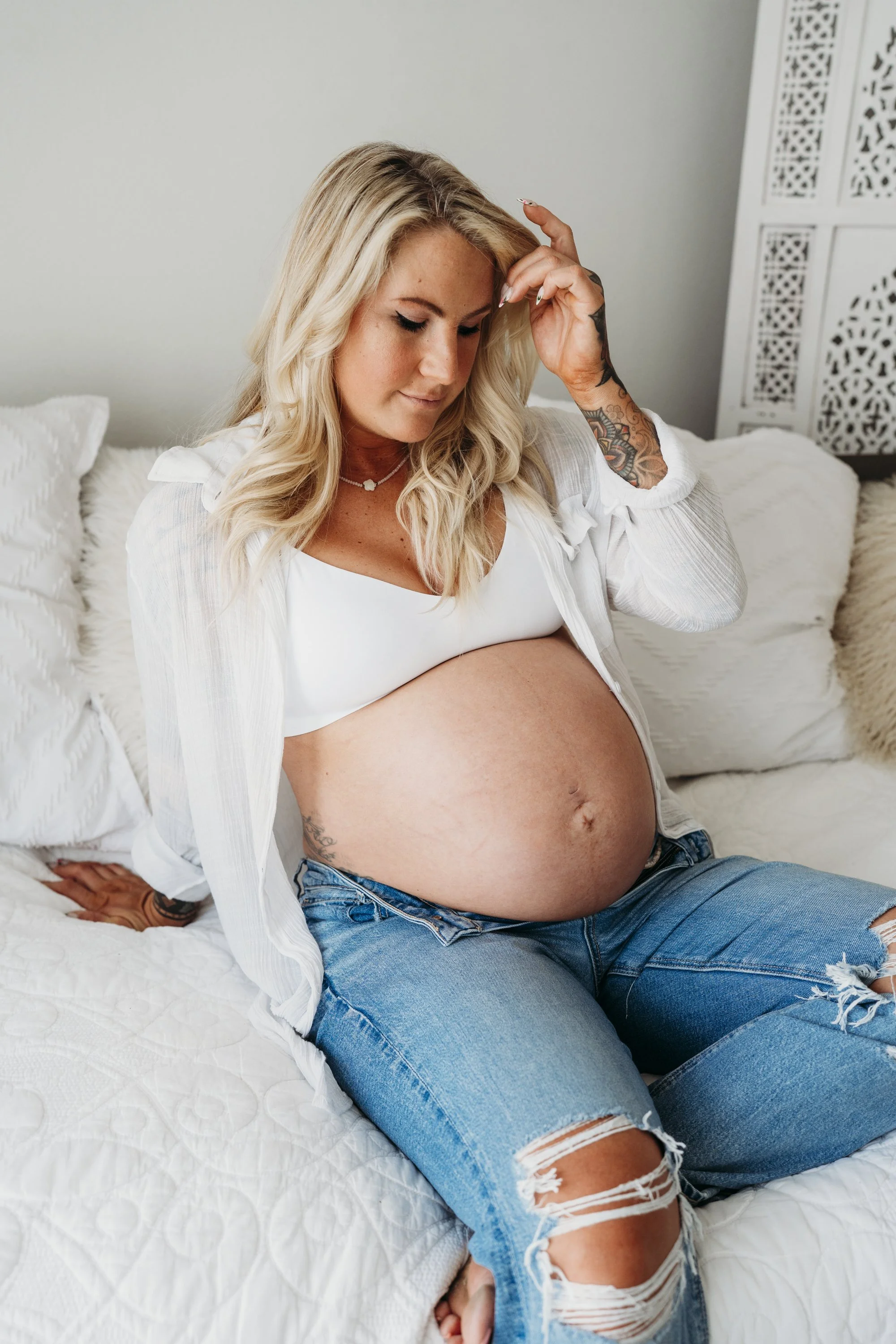 Pregnant woman sitting on bed, looking down, with blonde hair, tattoos, wearing a white top and ripped jeans.