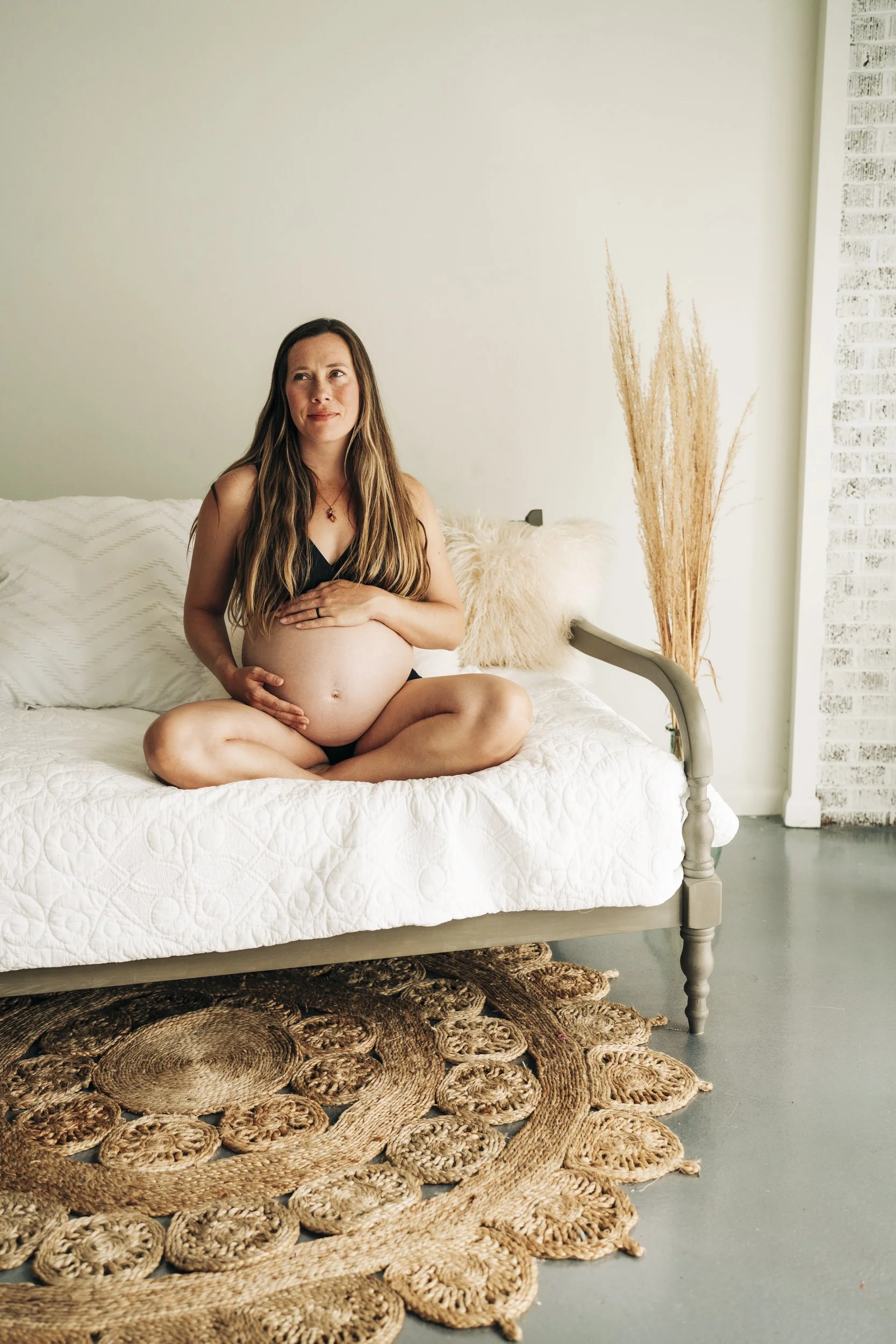 A pregnant woman sitting cross-legged on a bed with white bedding, holding her belly and looking thoughtfully to the side in a minimalist room with a rug and tall dried grasses.