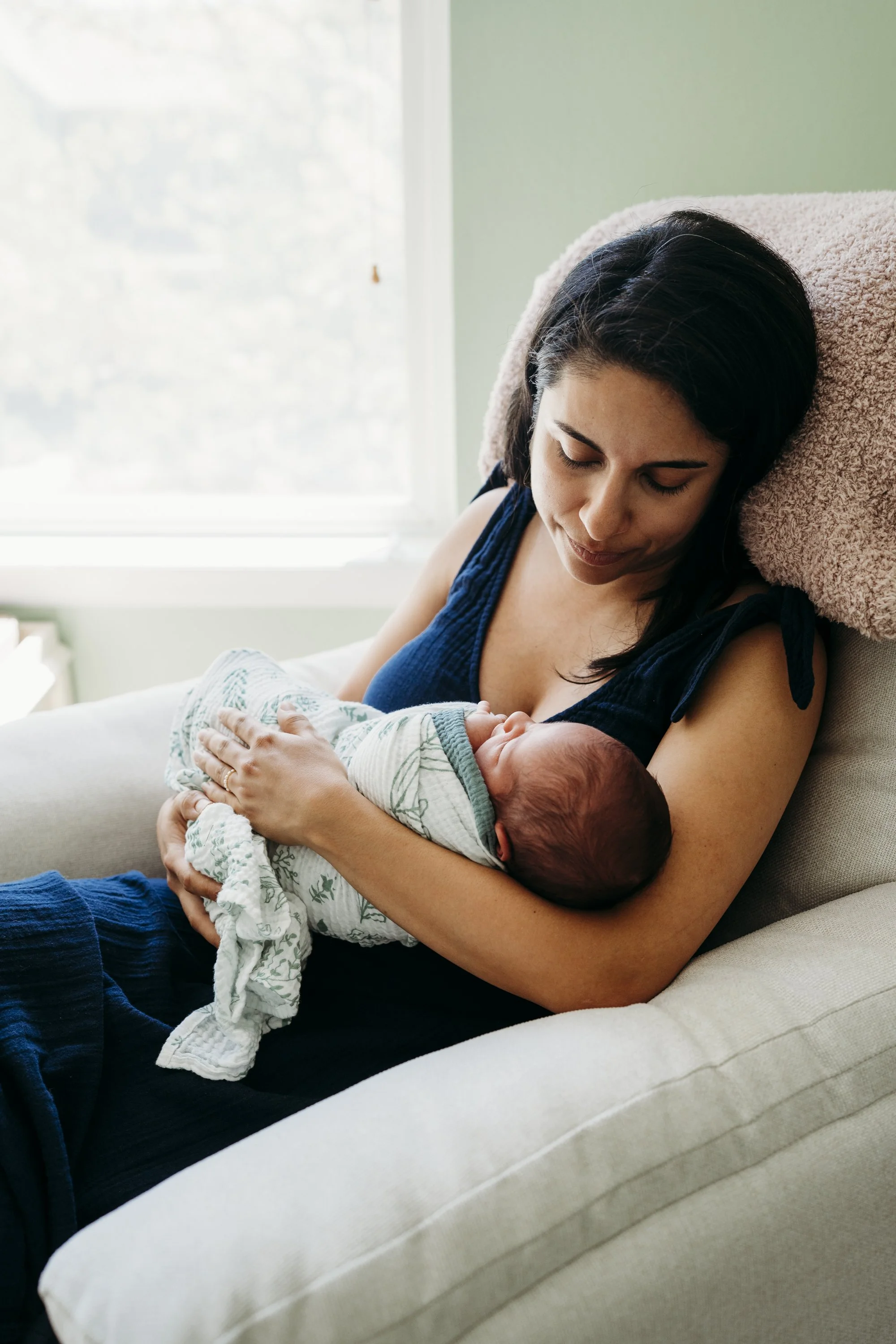 Woman holding a newborn baby on a sofa, near a window.