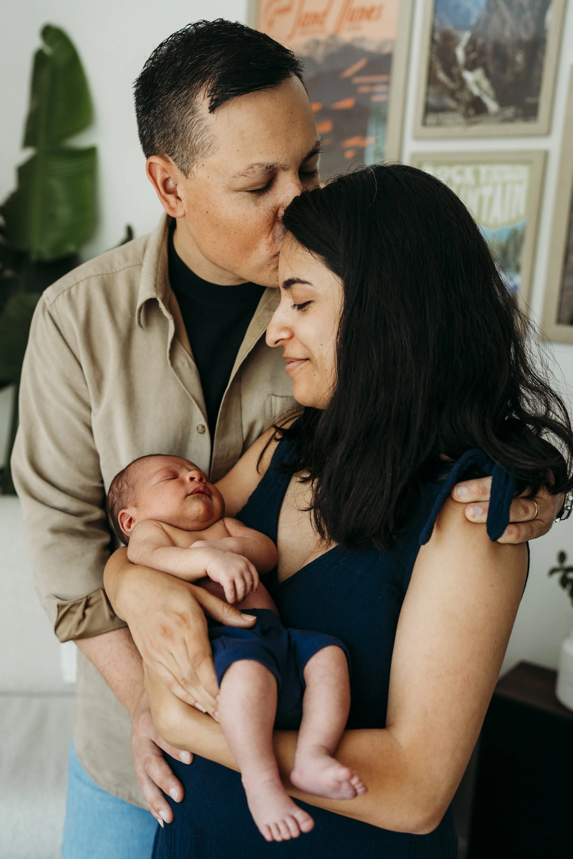 A couple holding their newborn baby, with the father kissing the mother's forehead, all smiling and sharing an intimate moment.