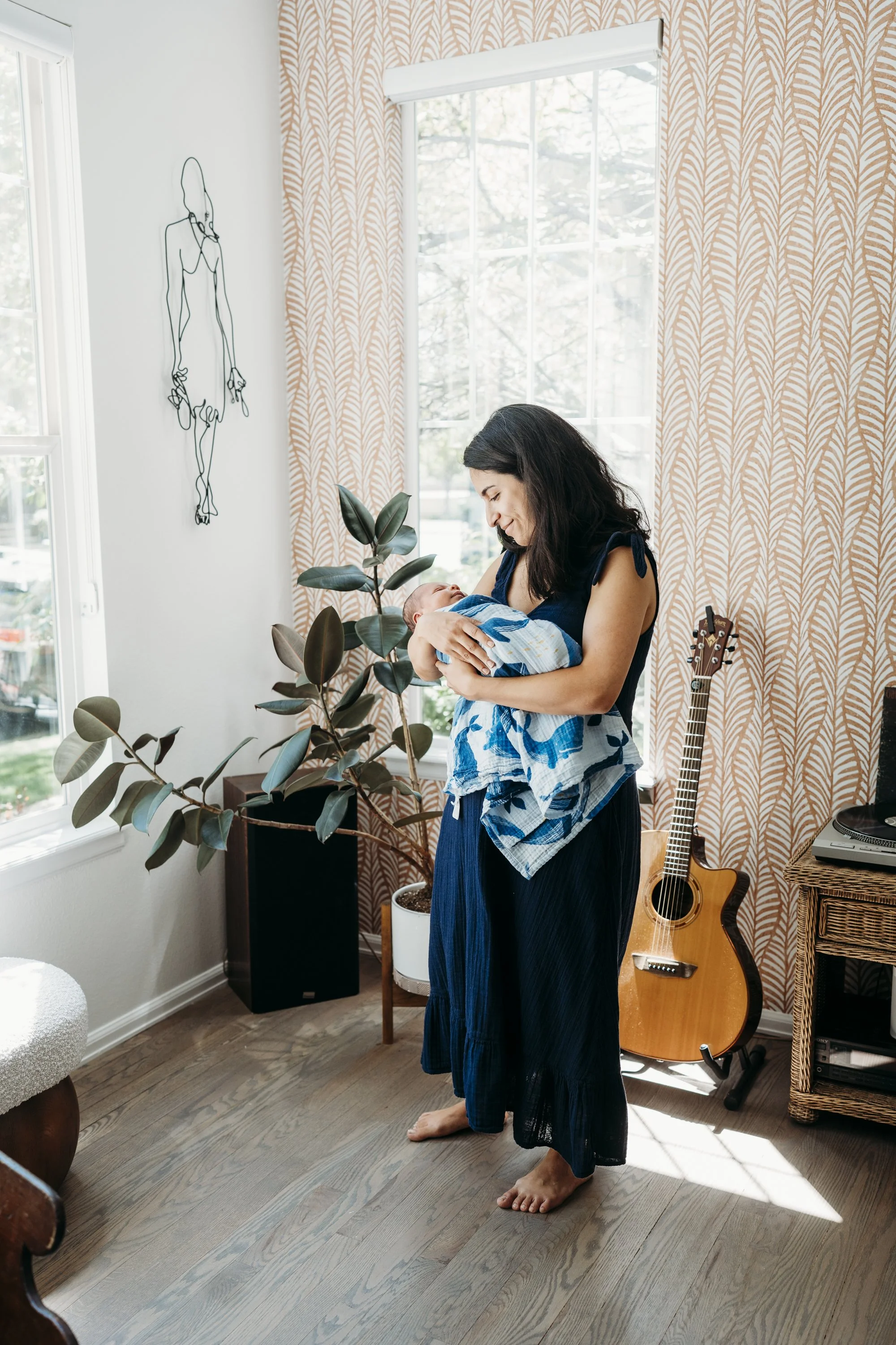 A woman holding a baby in a sunlit room with patterned curtains and a large plant, standing barefoot on wooden floor, near an acoustic guitar and digital piano.