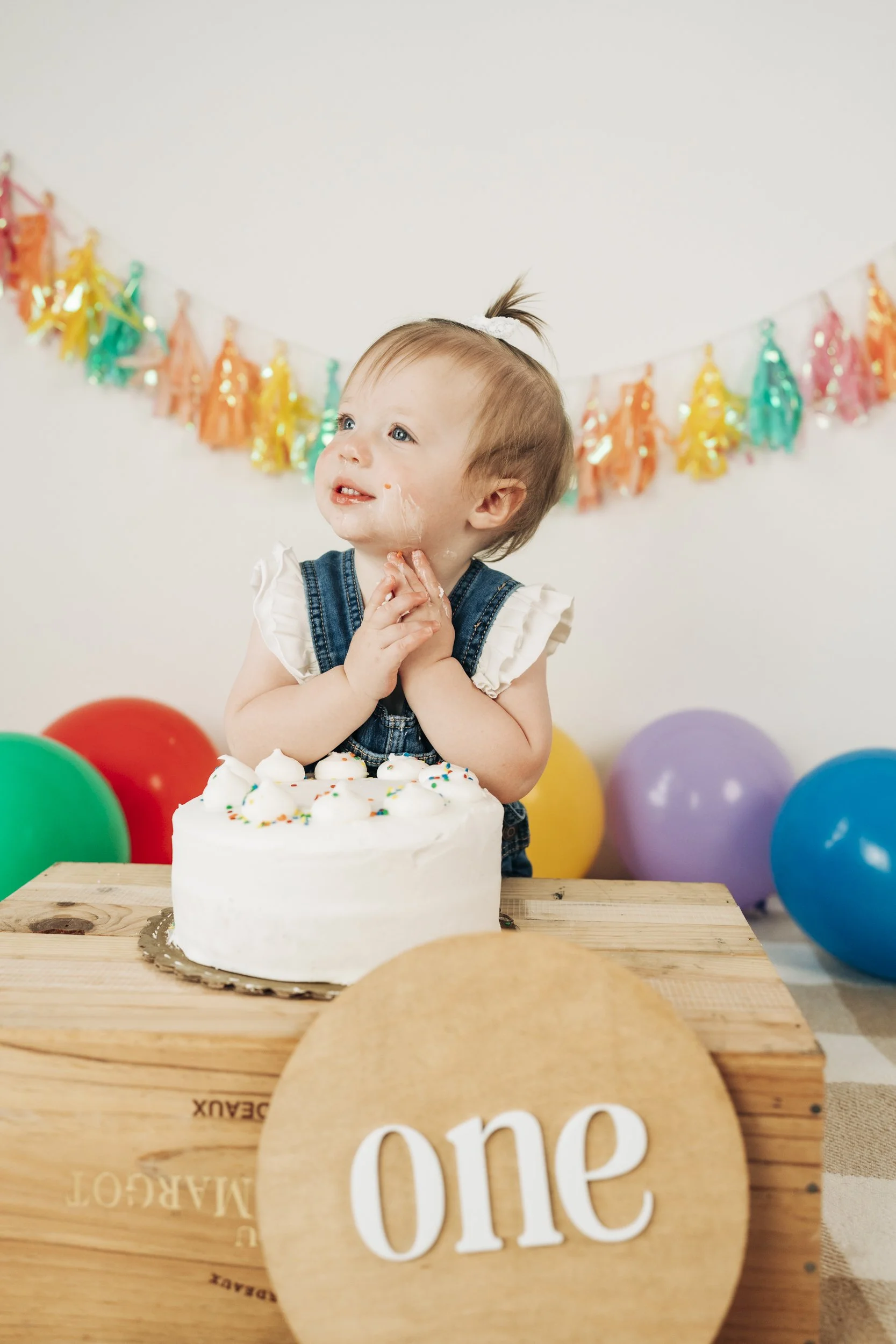 A young girl celebrating her first birthday with a cake, balloons, and colorful decorations in the background.