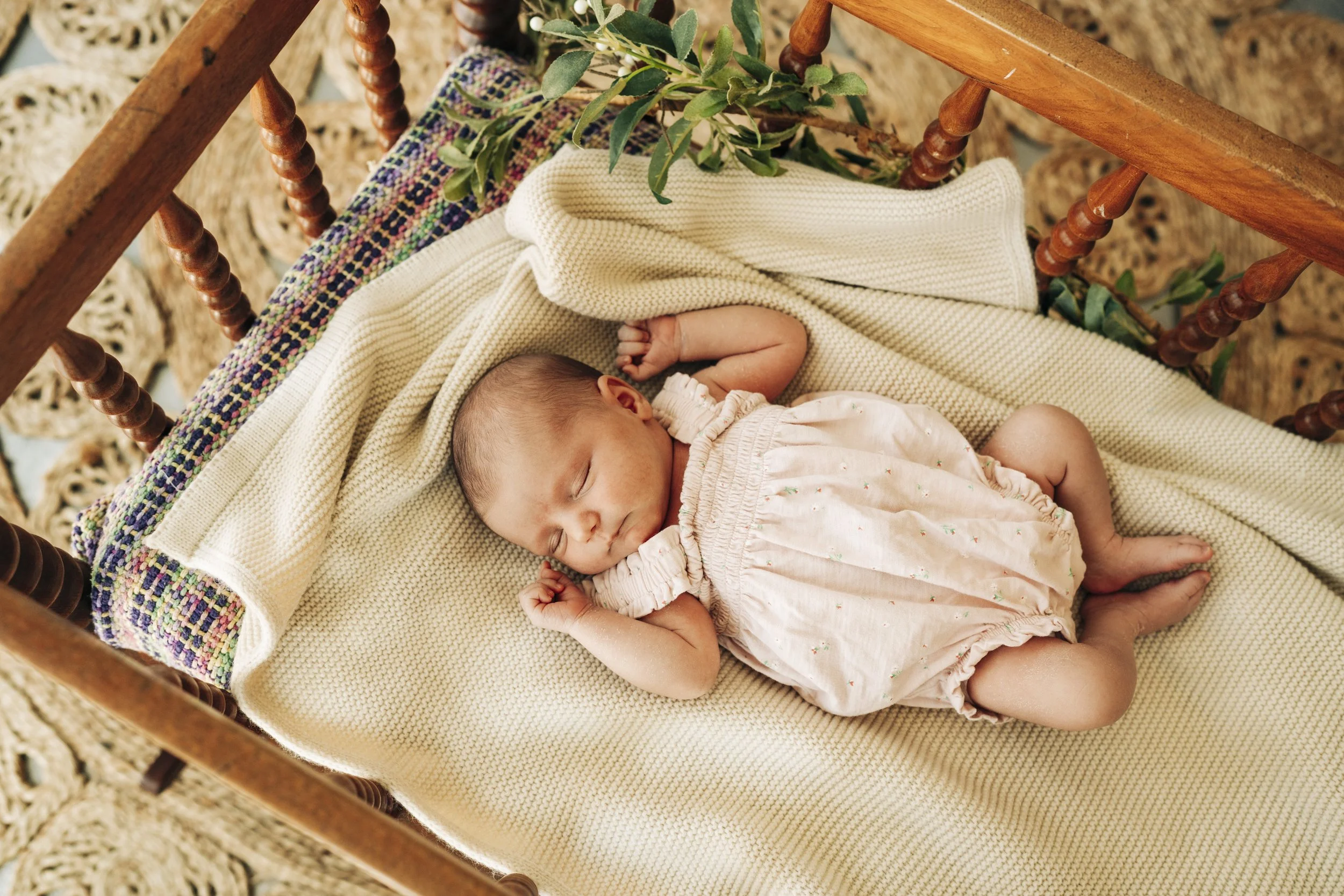 A sleeping baby girl lying on a cream blanket inside a wooden cradle, with a knitted cardigan and a small green plant nearby.