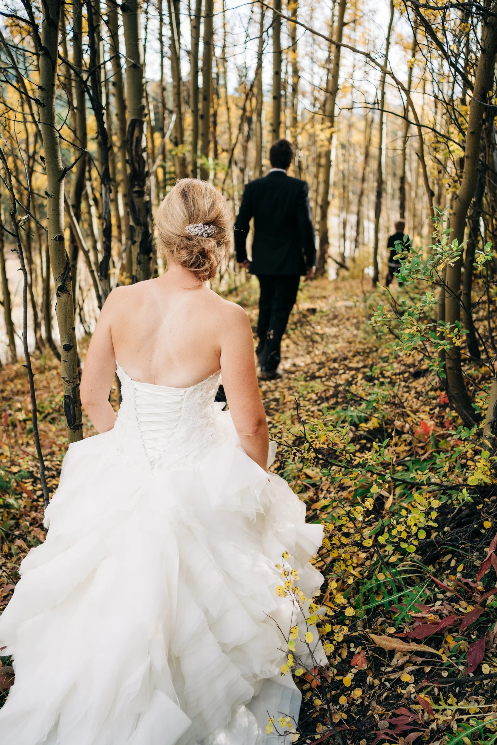 A bride in a white wedding dress with an elaborate train, sitting on the ground in a wooded area, with a man in a dark suit walking away in the background.