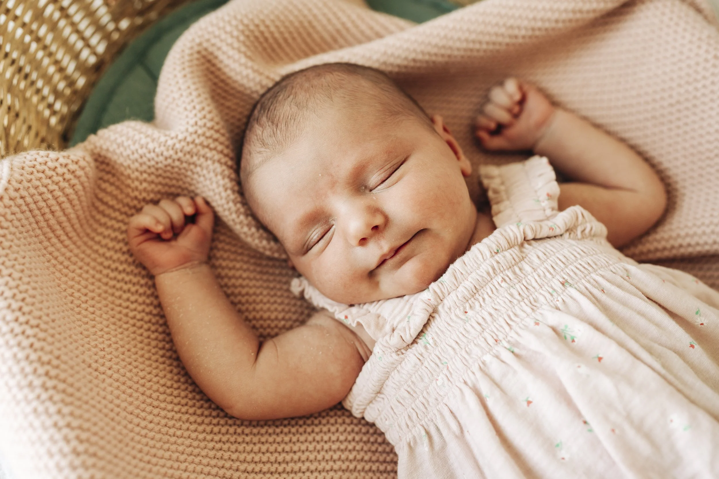 A smiling baby girl sleeping, lying on a pink knitted blanket with a beige dress and a pink hat.