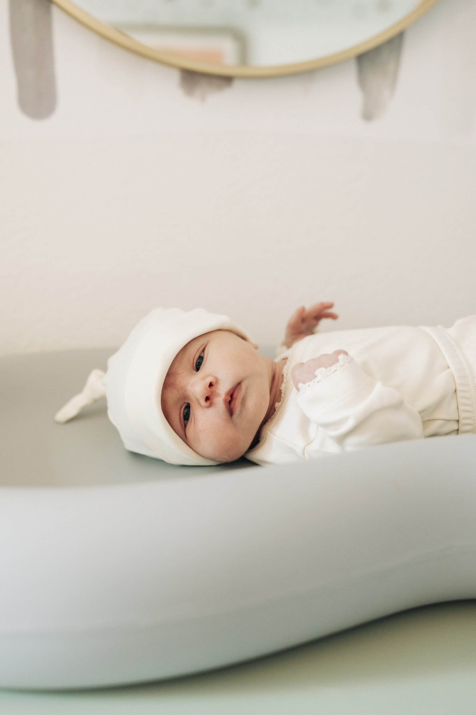 Newborn baby lying in a white crib, wearing a white hat and outfit, looking at the camera.