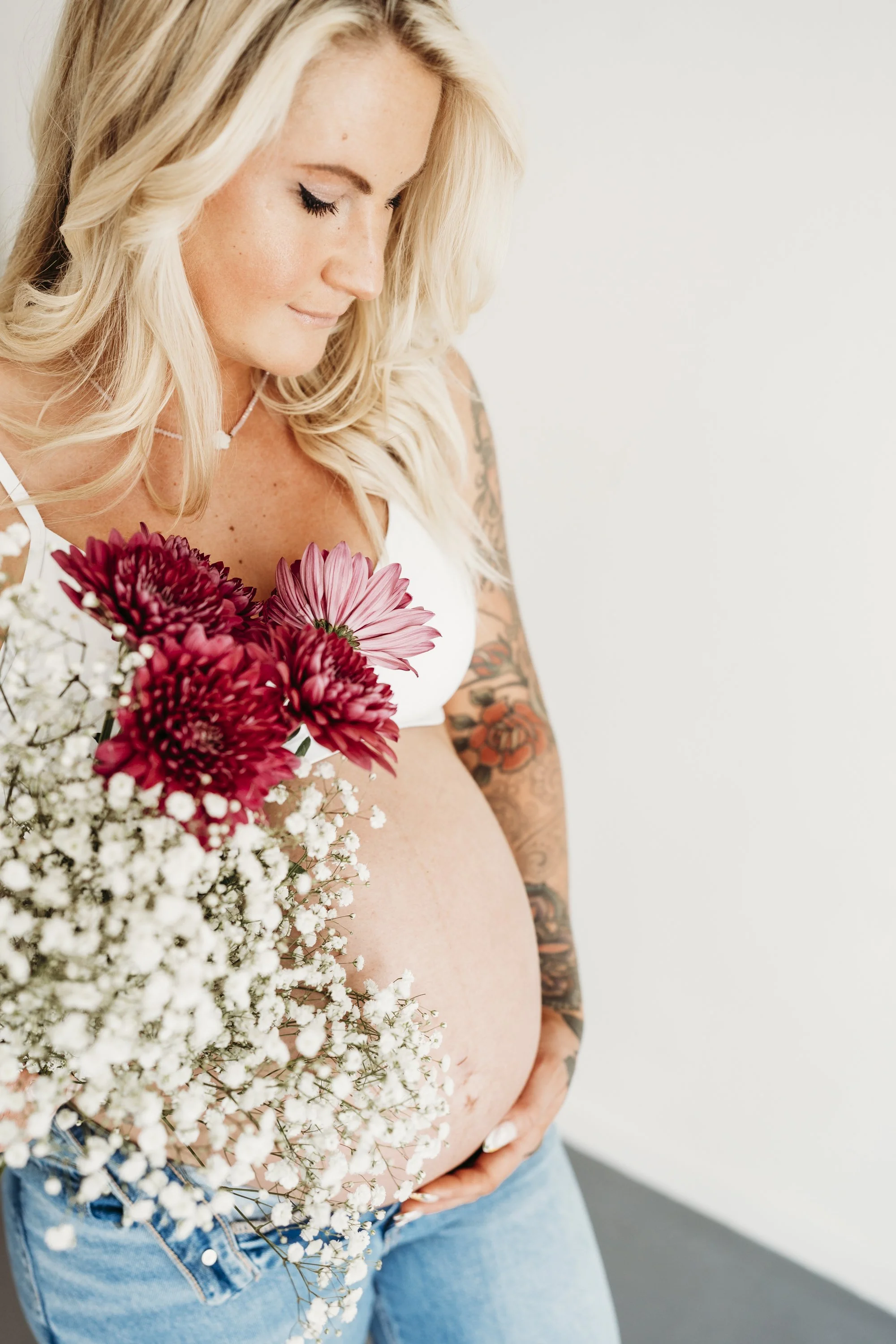 Pregnant woman with tattoos holding a bouquet of pink and white flowers, wearing a white top and jeans, standing against a white background.