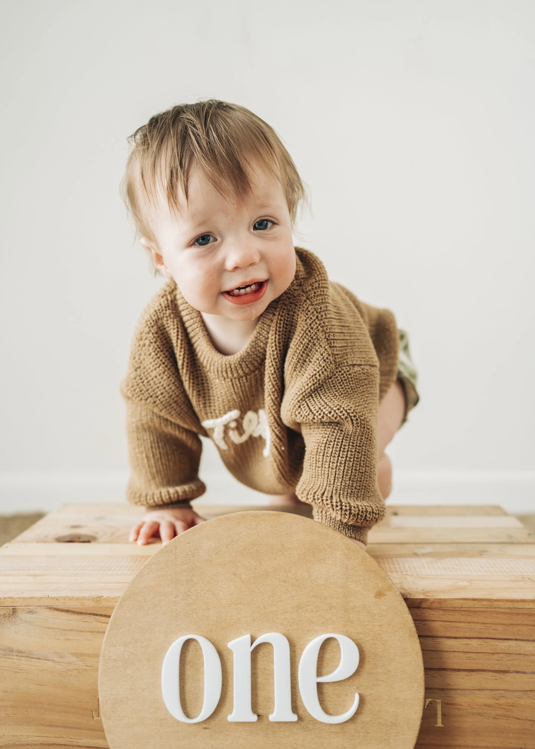 A young child with light brown hair and blue eyes on all fours, smiling, wearing a tan knit sweater with white lettering, in front of a round sign that says 'one', with a wooden surface.
