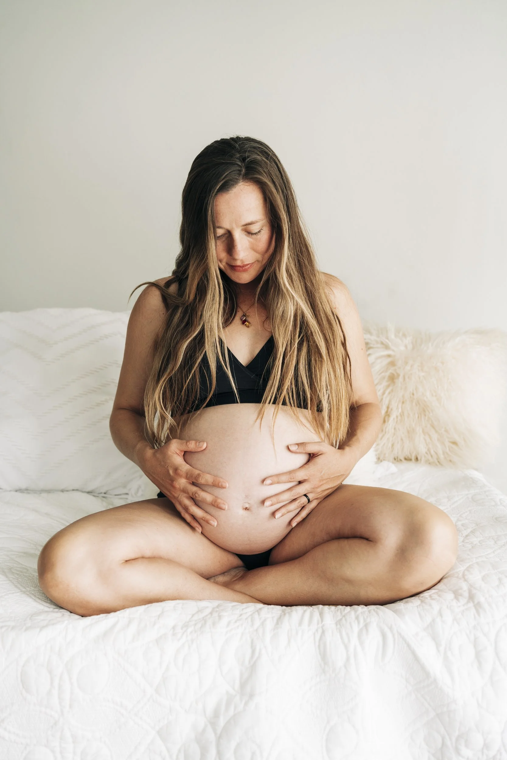 Pregnant woman sitting cross-legged on a bed, holding her belly and smiling gently.