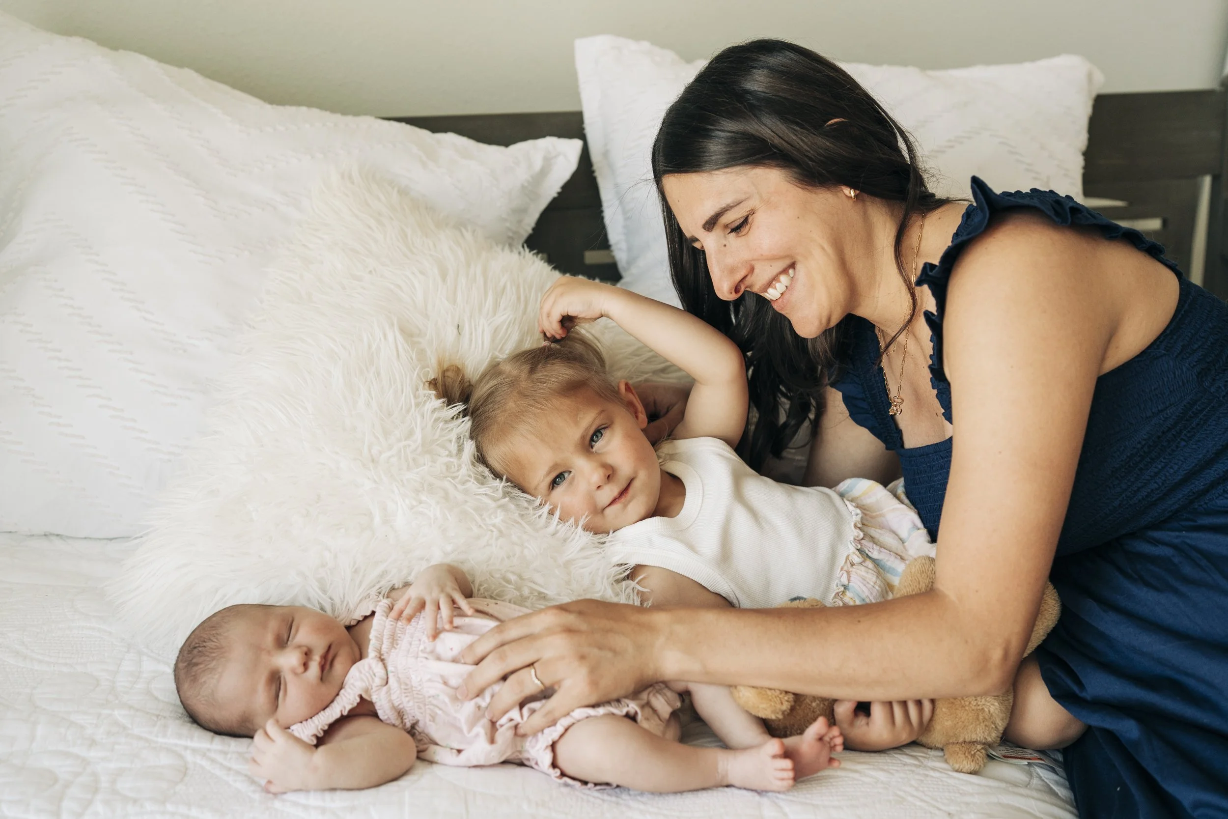 A woman with dark hair smiling and playing with two children, a toddler girl and a baby, on a bed with white pillows and a furry pillow.