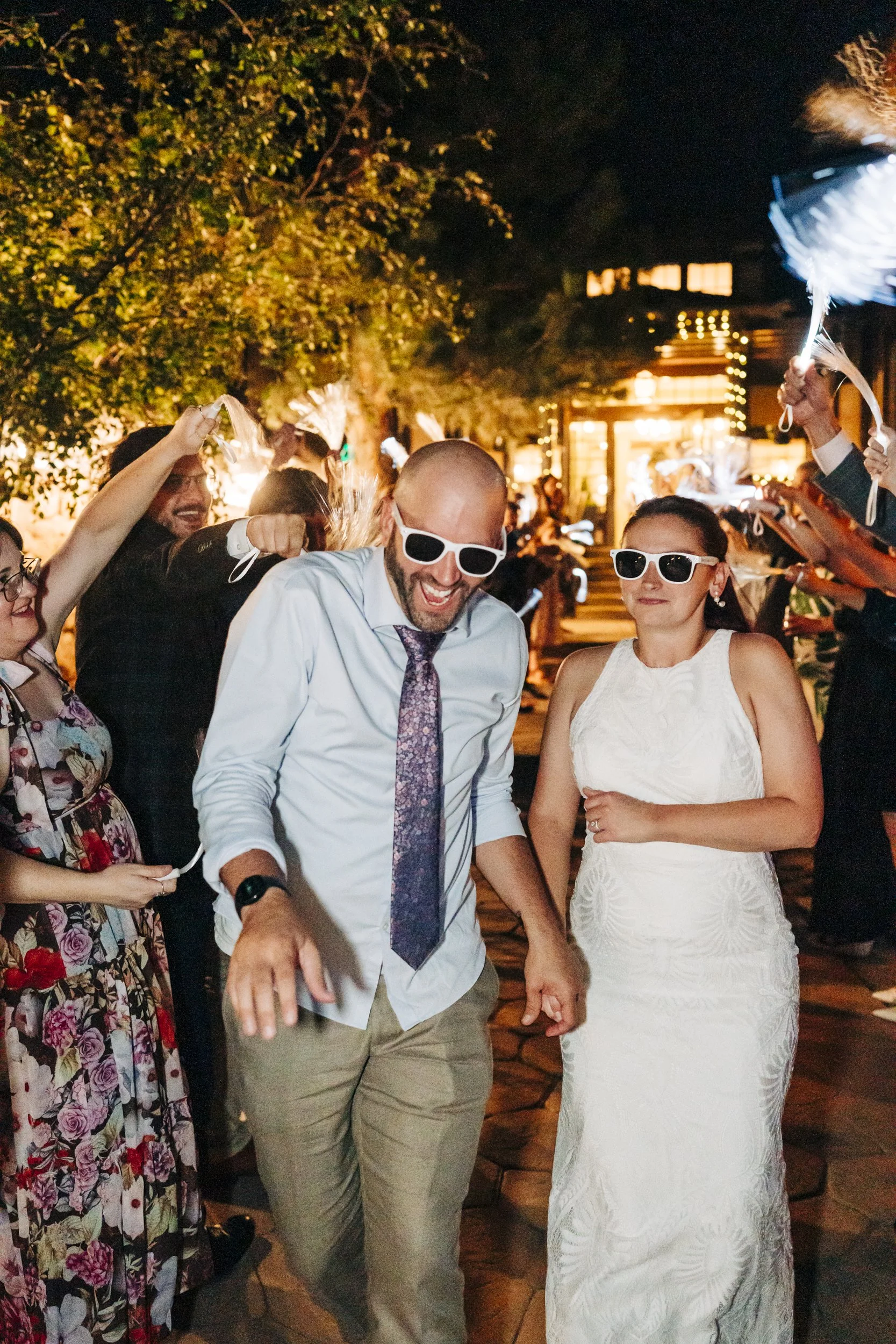 A group of people celebrating at night outdoors, with two women and a man in the center wearing sunglasses and smiling, surrounded by others holding sparklers. The scene is festive with warm lighting and trees in the background.