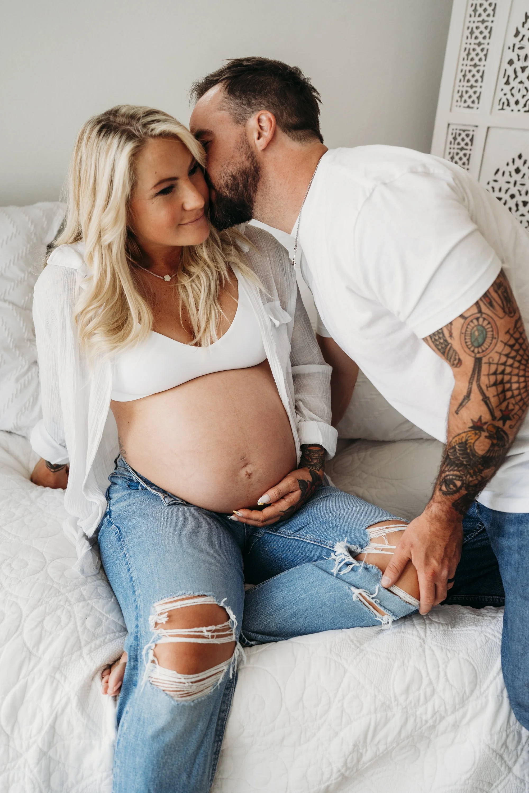 A pregnant woman with blonde hair in ripped jeans and a white bra, sitting on a bed. A man with tattoos, wearing a white t-shirt, leans in to kiss her on the forehead, holding her foot.