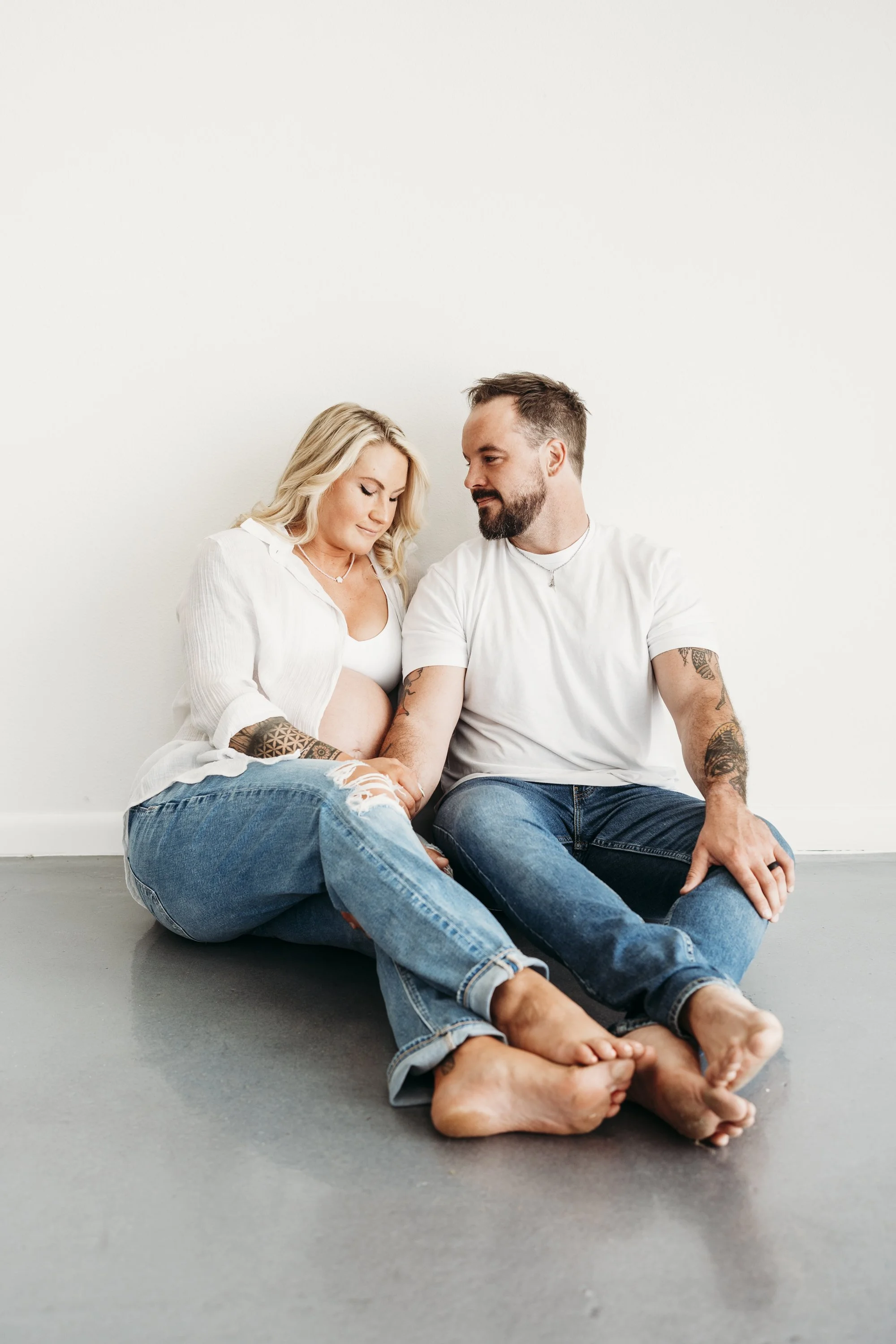 A pregnant woman and her partner sitting on the floor, holding hands and looking lovingly at each other against a plain white wall.